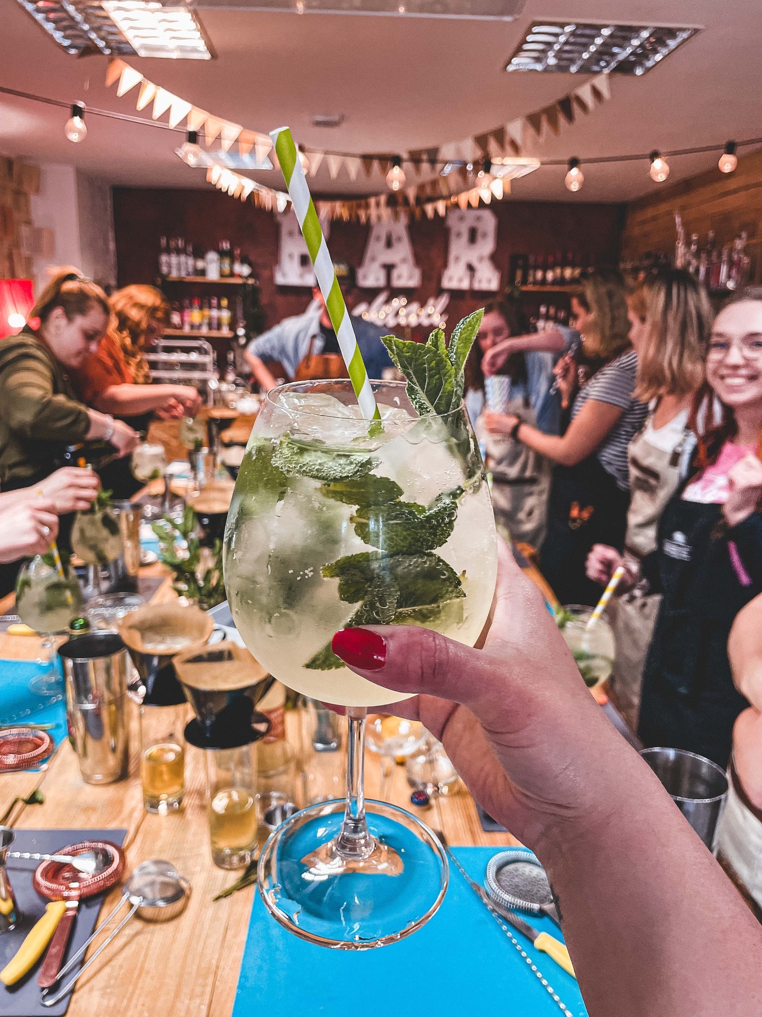 Warsztaty barmańskie: dłoń trzyma drinka Mojito na tle grupy osób przygotowujących koktajle, dekoracje i barowe akcesoria na drewnianym stole.