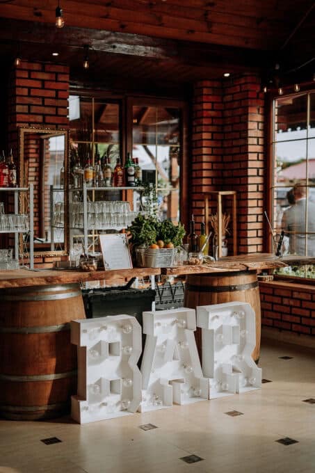 Rustykalny bar z drewnianym blatem na beczkach, podświetlonym napisem 'BAR' i dekoracjami roślinnymi w ceglanym wnętrzu.