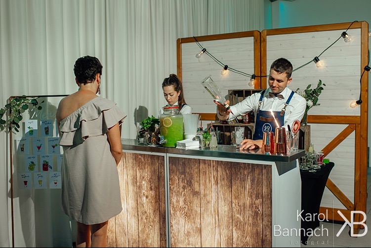 Barman w akcji przygotowuje drinka dla klientki przy barze z menu koktajli. W tle dekoracja z żarówek i pomocniczka. Profesjonalny catering na imprezę.