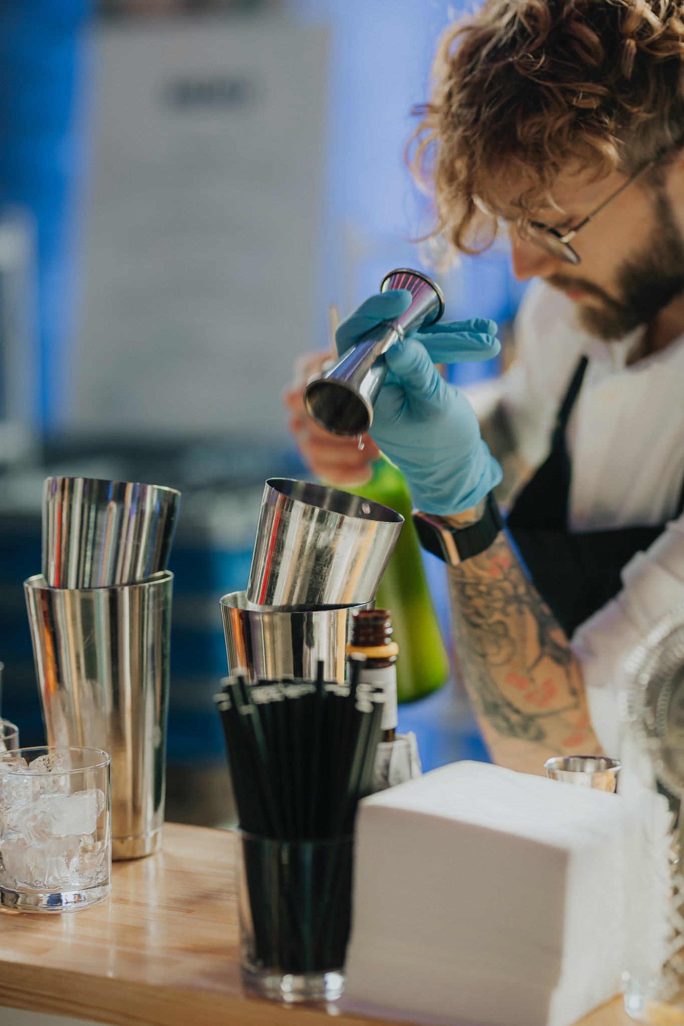 Barman w Gdańsku przygotowuje drinka, odmierzając składniki w miarce barmańskiej. Widoczne shakery, lód i inne akcesoria barmańskie.