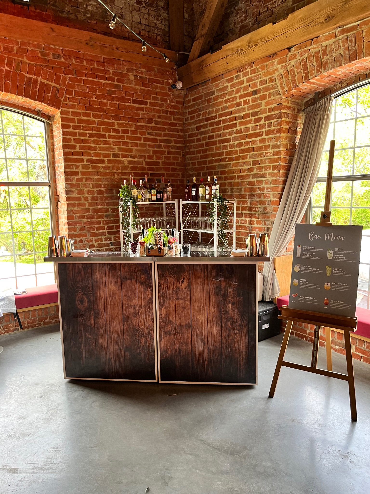 Mobilny bar z menu drinków na sztaludze, ustawiony w przestronnym wnętrzu z ceglanymi ścianami i dużymi oknami. Dekoracje roślinne i butelki na barze.