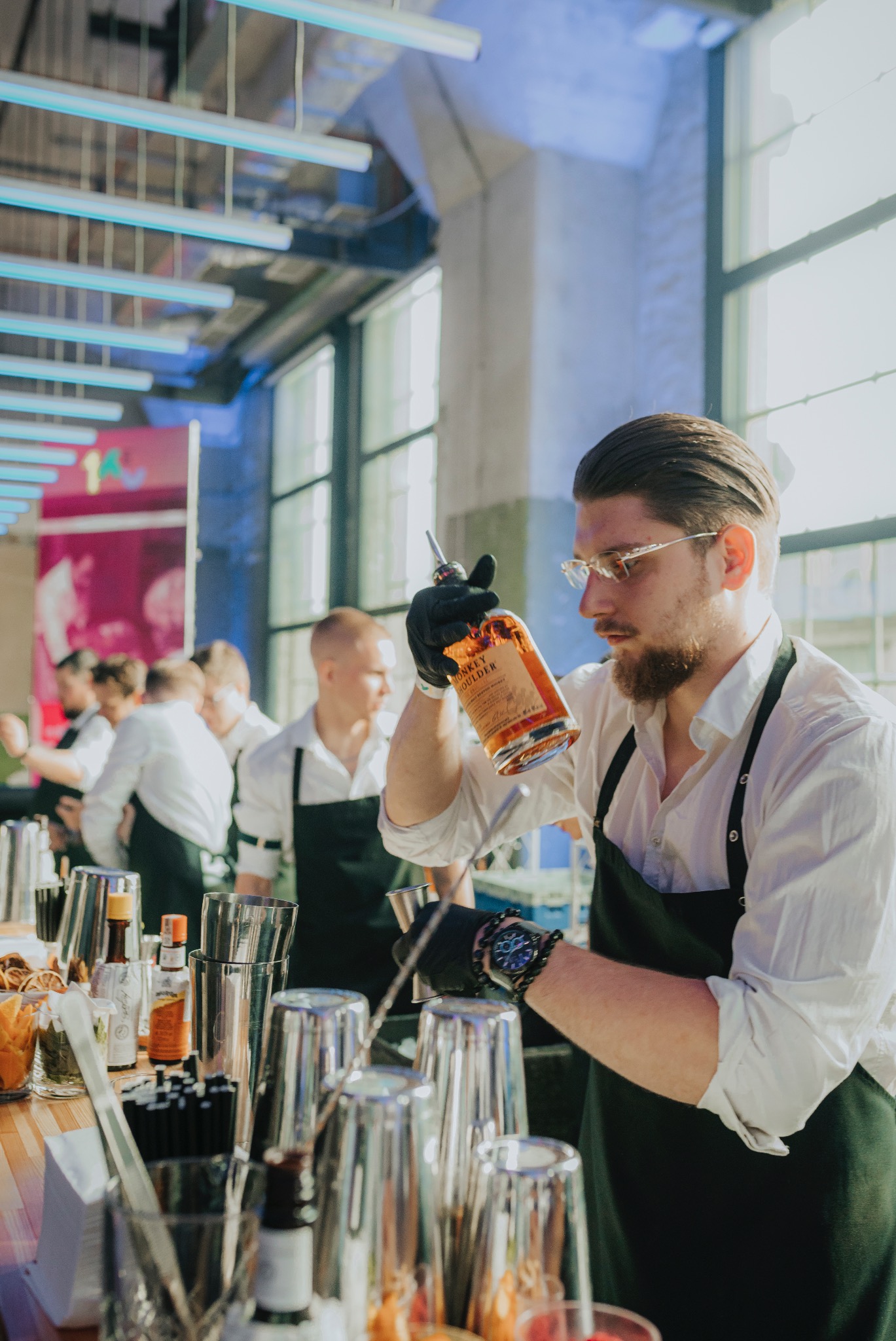 Barman w czarnych rękawiczkach nalewa whisky Monkey Shoulder do miarki, przygotowując koktajl na barze podczas eventu. W tle inni barmani i industrialne okna.