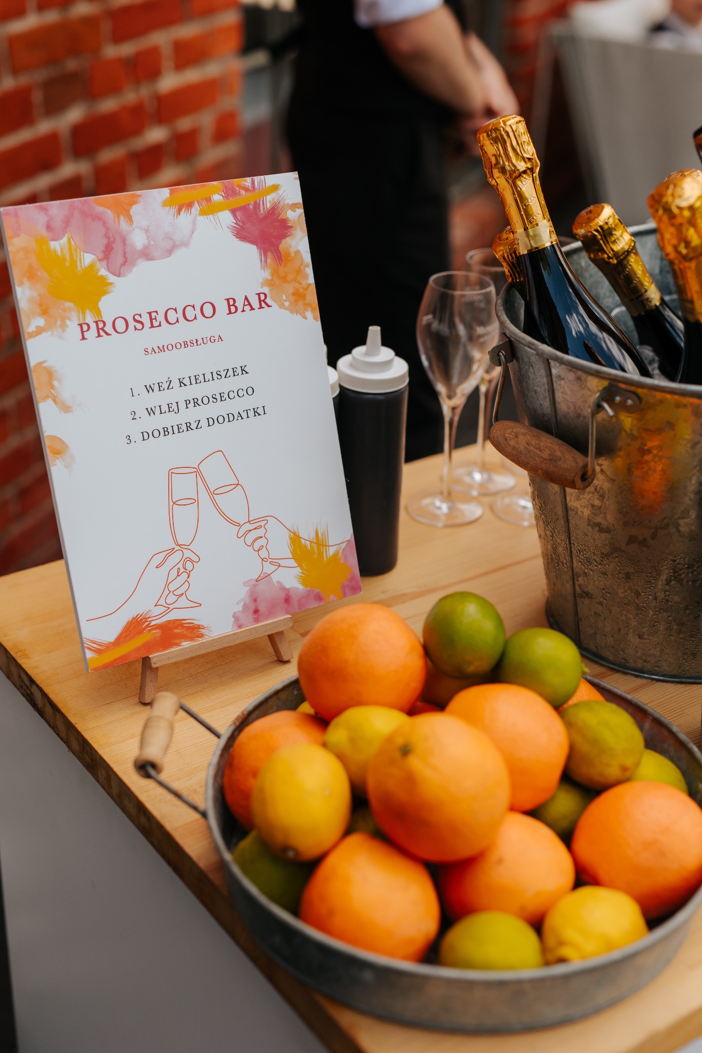 Stół z prosecco bar: instrukcja samoobsługi na sztaludze, wiadro z butelkami i owocami. Element dekoracji eventowej, detale przyjęcia.