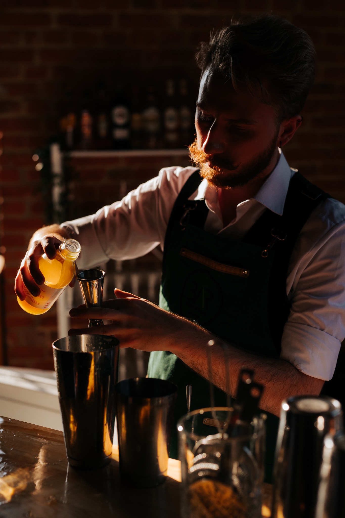 Barmen z rudą brodą w zielonym fartuchu nalewa żółty płyn z butelki do miarki nad metalowym shakerem. W tle ceglana ściana i butelki.