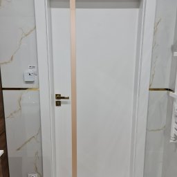 Montownia Mateusz Michalczuk - Białe drzwi wewnętrzne z pionowym, jasnym paskiem i złotą klamką, zamontowane w łazience z marmurowymi płytkami. Widoczny regulator temperatury i gniazdko.