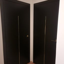 Montownia Mateusz Michalczuk - Dwa czarne, nowoczesne drzwi z złotymi akcentami, zamontowane w rogu pomieszczenia. Widoczne detale wykończenia i minimalistyczny design.