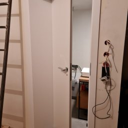 Montownia Mateusz Michalczuk - Białe, częściowo otwarte drzwi wewnętrzne z widocznym fragmentem łazienki w tle. Obok widoczne wystające kable elektryczne i drabina oparta o ścianę.