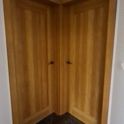 Montownia Mateusz Michalczuk - Dwa drewniane skrzydła drzwiowe, zamontowane w rogu pomieszczenia, z widoczną strukturą drewna i ciemną, marmurkową podłogą. Nietypowe rozwiązanie architektoniczne.