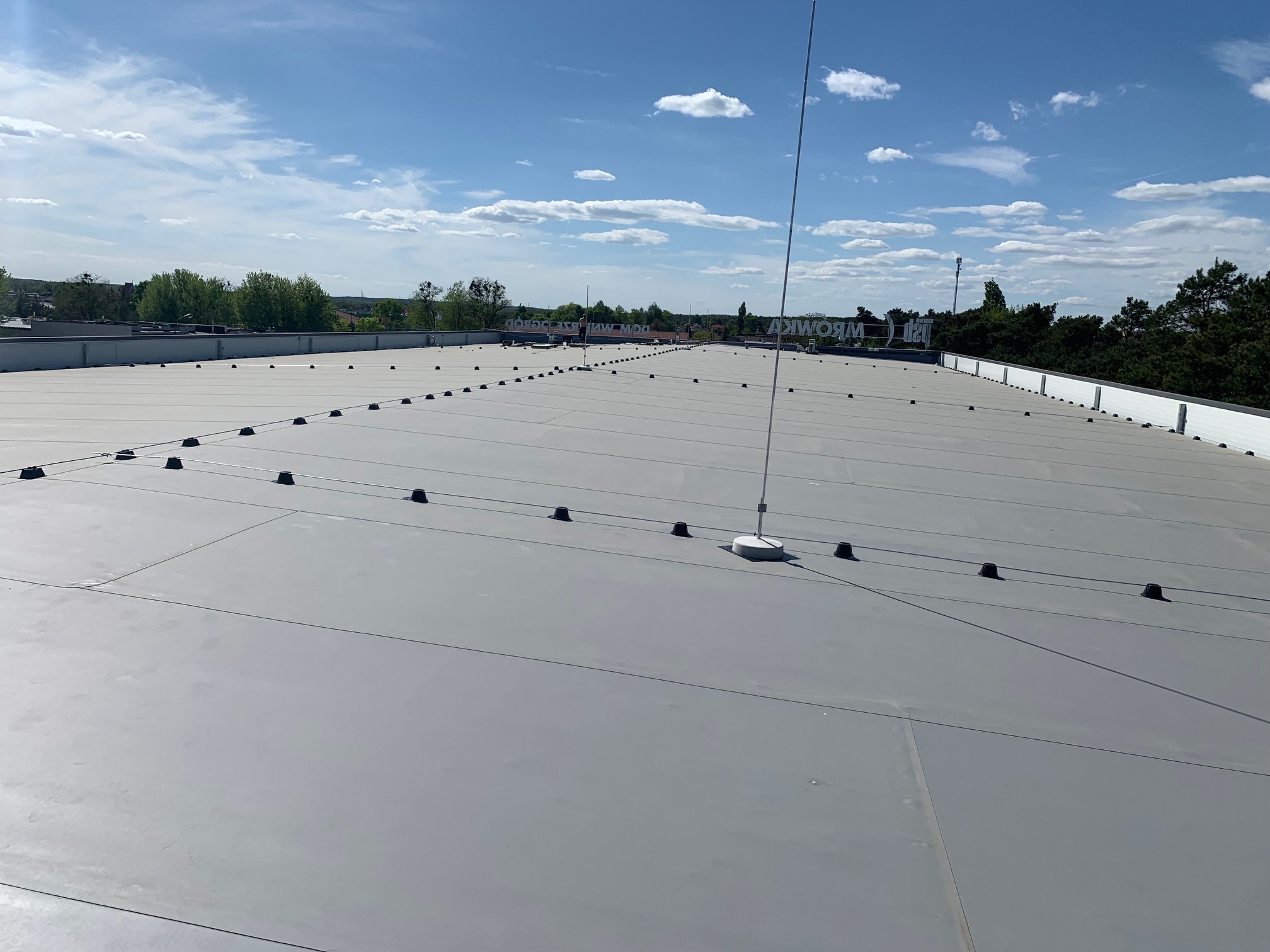 Szary dach przemysłowy z systemem odgromowym i elementami mocującymi, widok z góry w słoneczny dzień. Widoczne niebo z chmurami i budynki w tle.