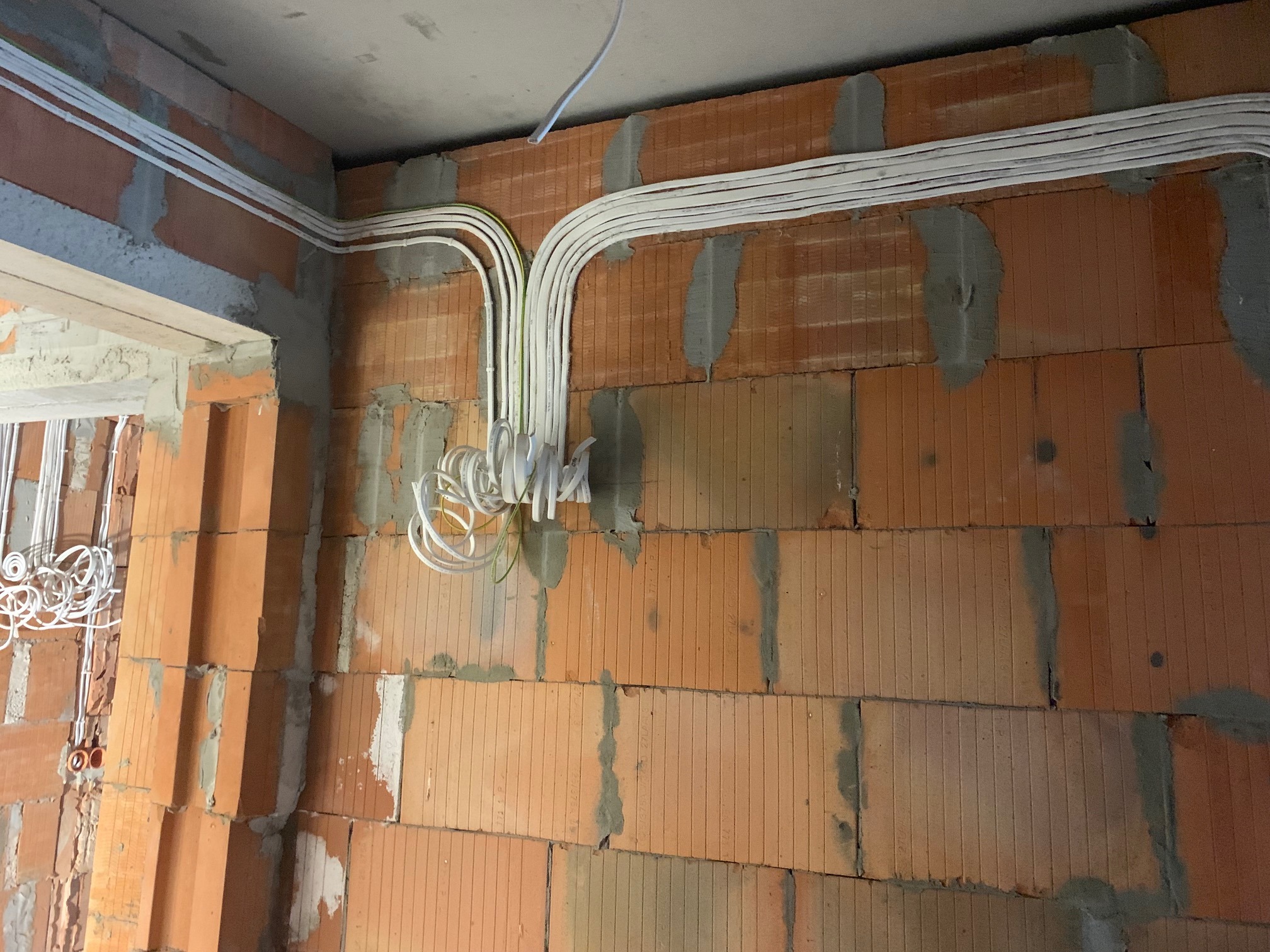 Białe przewody elektryczne ułożone wzdłuż ceglanej ściany w surowym stanie, widoczne łączenia i puszki instalacyjne. Instalacja w trakcie montażu.