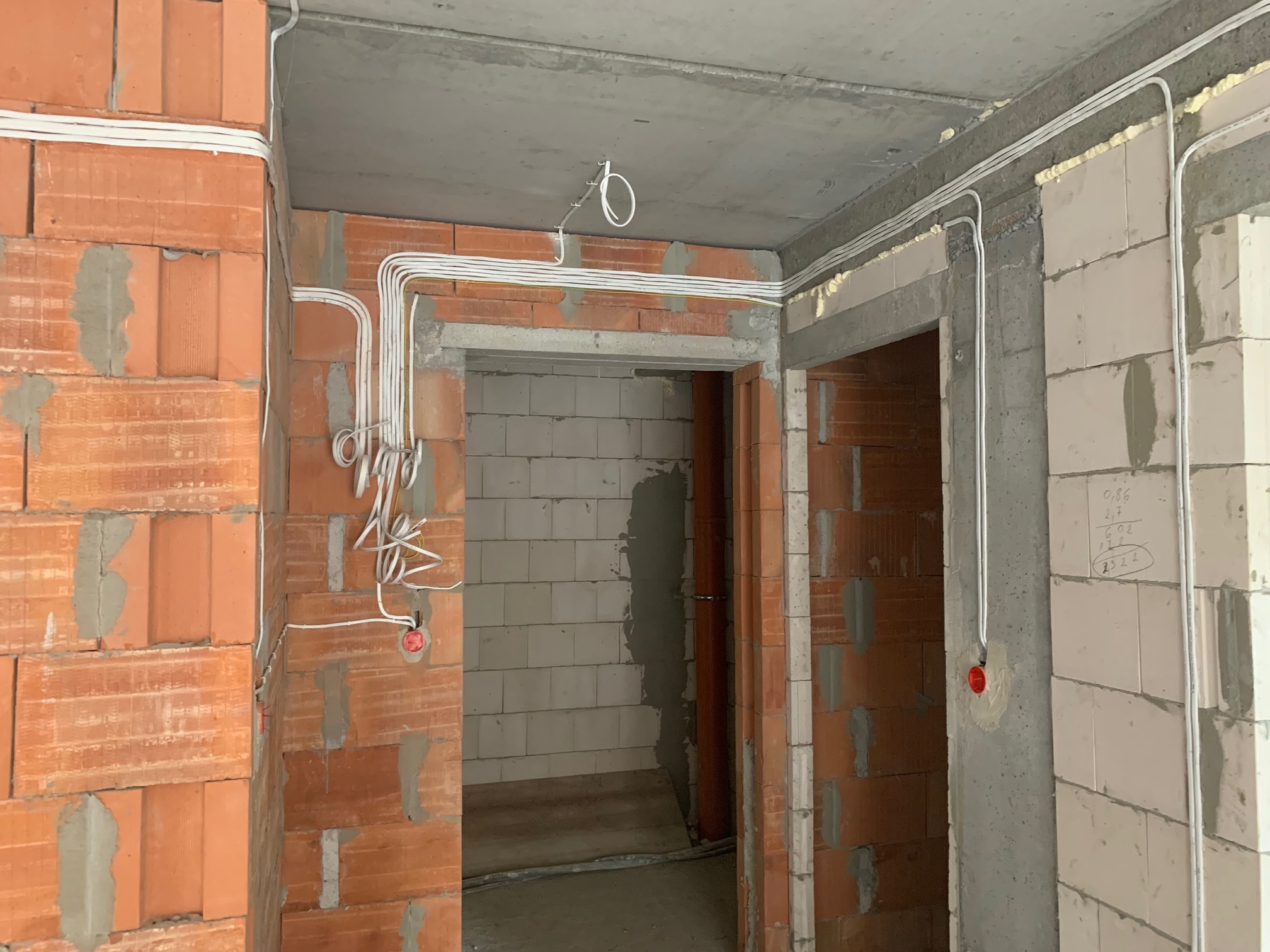 Białe przewody elektryczne ułożone wzdłuż surowych ścian z czerwonej cegły i betonu wewnątrz budynku w trakcie budowy. Widoczne puszki instalacyjne.