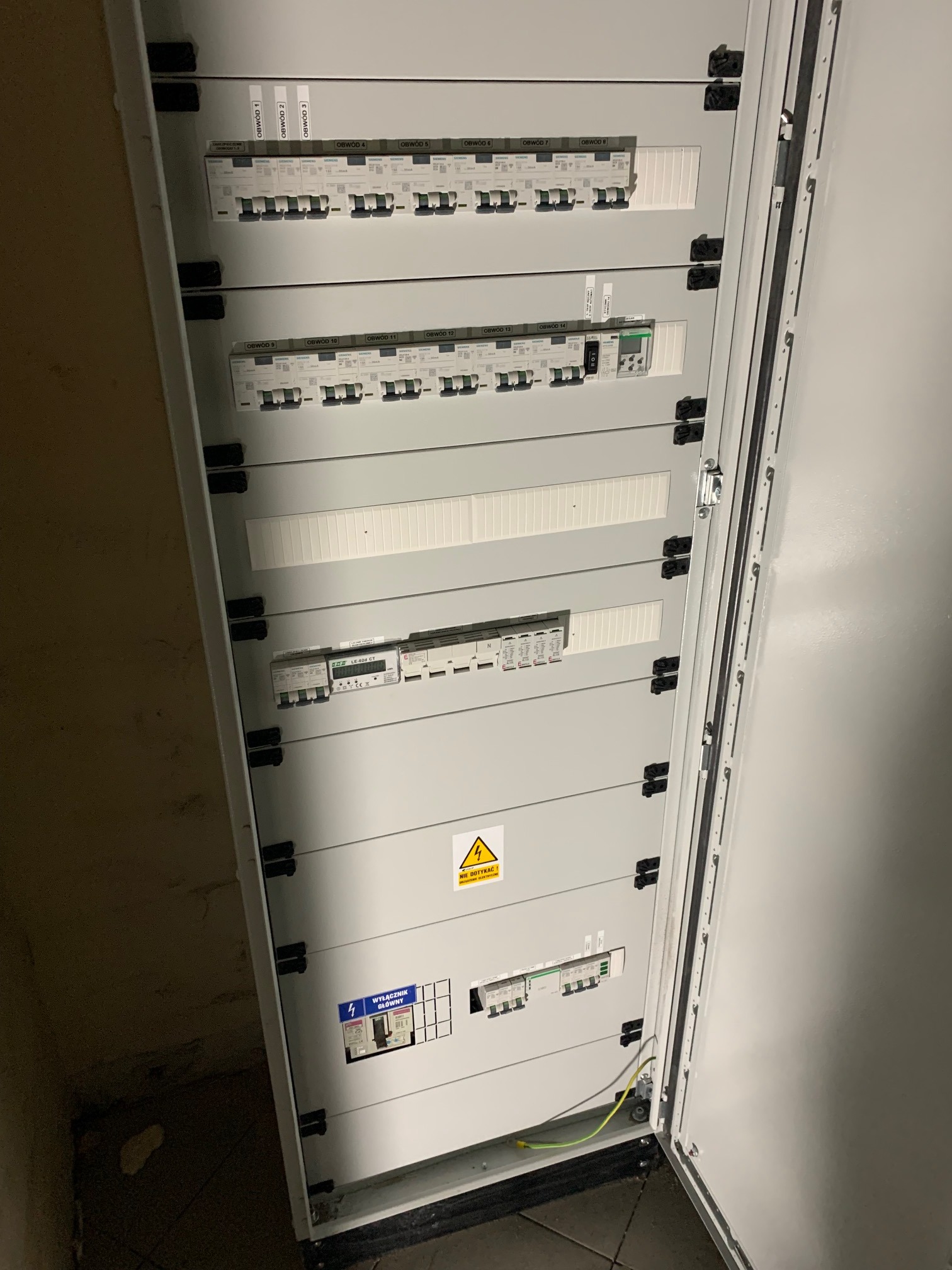 Otwarta szafa elektryczna z bezpiecznikami, opisana obwodami, z żółtym znakiem ostrzegawczym 'Nie dotykać', widoczny przewód uziemiający.