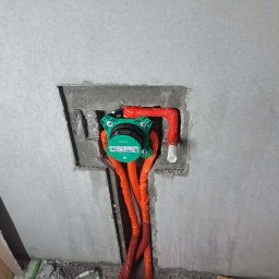 AquaMaster Usługi Hydrauliczne - Zielony podtynkowy zestaw prysznicowy Hansgrohe z pomarańczowymi rurami w surowej, szarej ścianie łazienki, widoczne elementy instalacji wodnej.