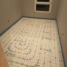 AquaMaster Usługi Hydrauliczne - Biała mata grzewcza z rurkami w kształcie pętli, zamocowanymi czerwonymi i czarnymi uchwytami, ułożona na podłodze w pomieszczeniu z oknem dachowym, przygotowana do wylewki.