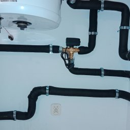 AquaMaster Usługi Hydrauliczne - Zamontowany bojler Galmet z izolowanymi rurami i zaworem trójdrożnym na białej ścianie z gniazdkiem elektrycznym i szarą glazurą podłogową. Instalacja wodna w łazience.