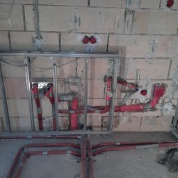 AquaMaster Usługi Hydrauliczne - Instalacja wodno-kanalizacyjna w trakcie montażu w surowej łazience. Widoczne czerwone rury, metalowa konstrukcja pod zabudowę i ściany z betonu komórkowego.