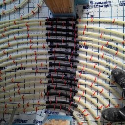 AquaMaster Usługi Hydrauliczne - Instalacja ogrzewania podłogowego z rur PEX mocowanych czerwonymi klipsami do płyty izolacyjnej z nadrukowaną siatką. Widoczne czarne, karbowane rury rozdzielacza.