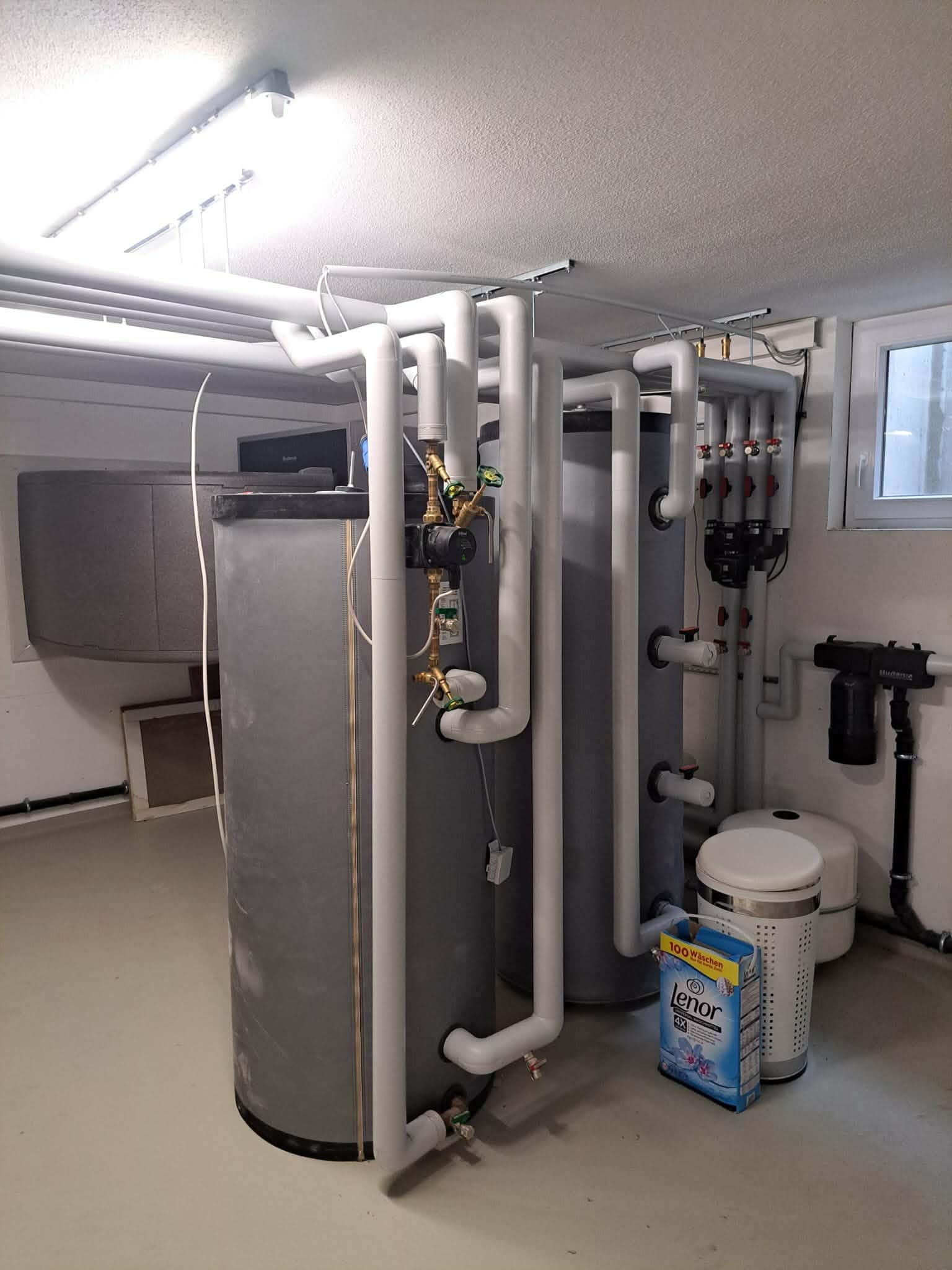 Instalacja grzewcza z trzema zbiornikami i rurami w piwnicy, widoczny filtr wody i zmiękczacz. Na podłodze stoi proszek do prania.