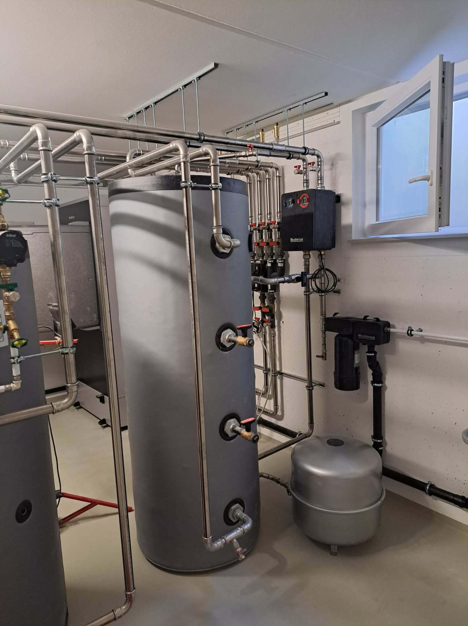 Nowoczesna kotłownia z dwoma szarymi zbiornikami, systemem rur ze stali nierdzewnej i czarnym panelem sterowania Buderus, oświetlona przez okno.