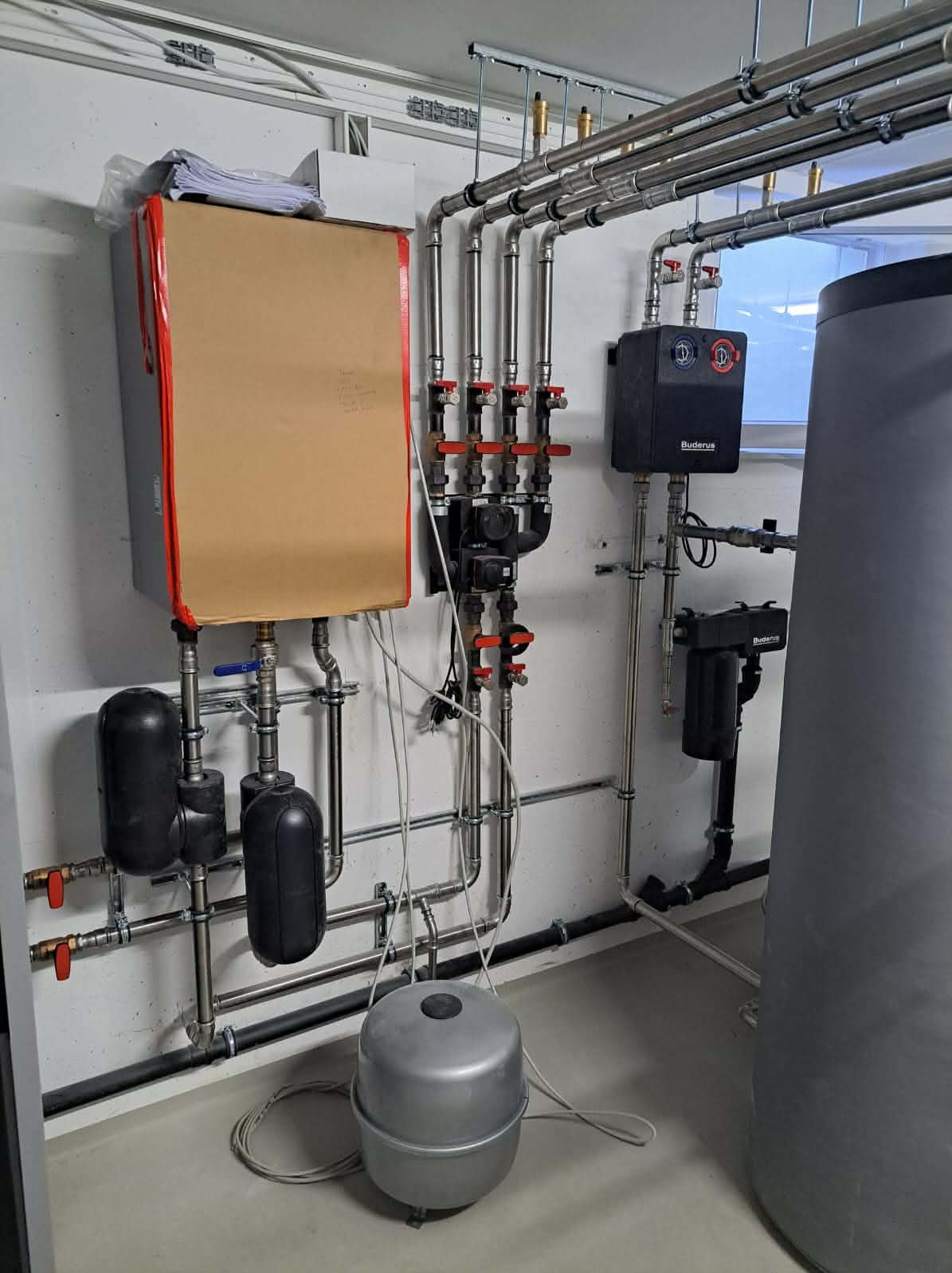 Instalacja grzewcza z kotłem, zbiornikami i rurami, widoczne zawory i czujniki. Kocioł częściowo zakryty kartonem. Elementy zamontowane na ścianie w pomieszczeniu.