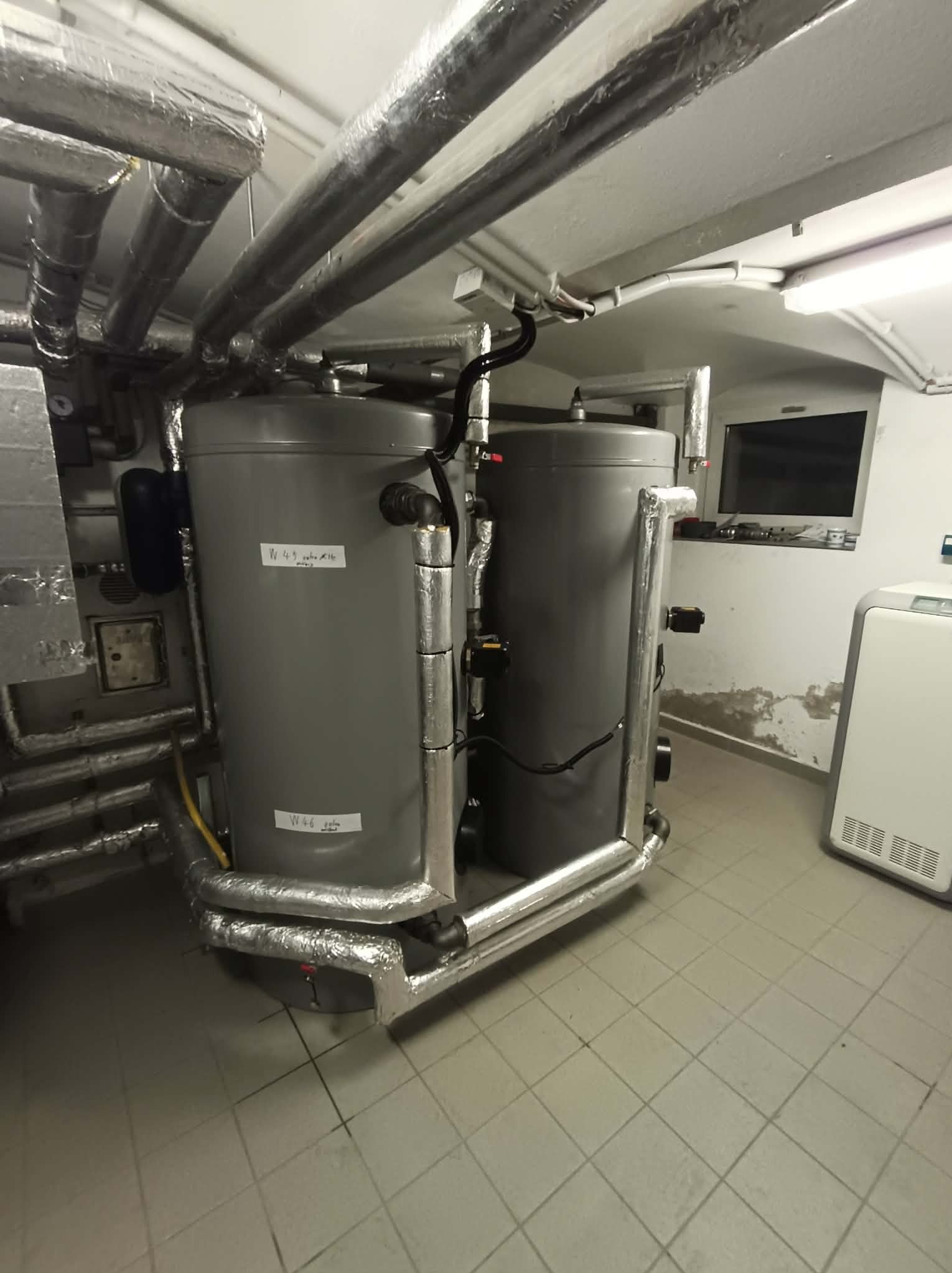 Dwa szare zbiorniki na wodę, połączone rurami z izolacją termiczną, w pomieszczeniu z kafelkami i oświetleniem sufitowym. Widoczne okno i białe urządzenie grzewcze.