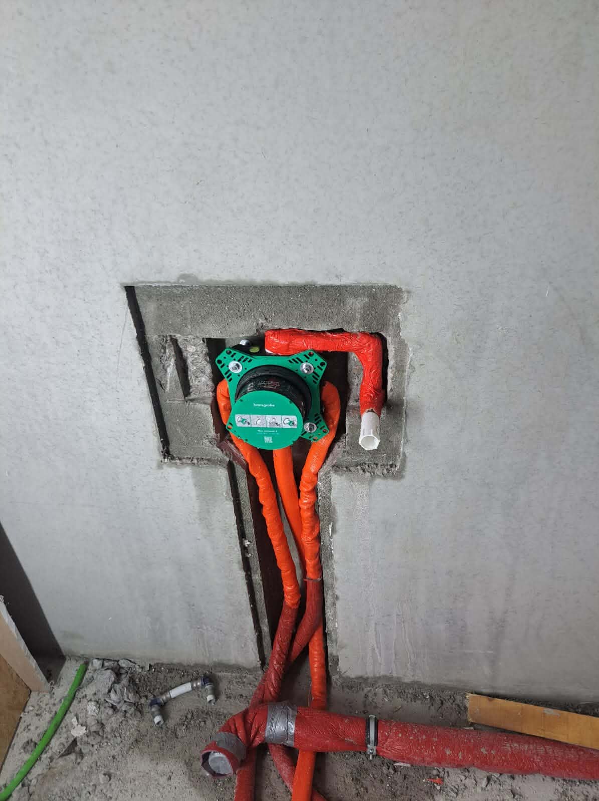 Zielony podtynkowy zestaw prysznicowy Hansgrohe z pomarańczowymi rurami w surowej, szarej ścianie łazienki, widoczne elementy instalacji wodnej.