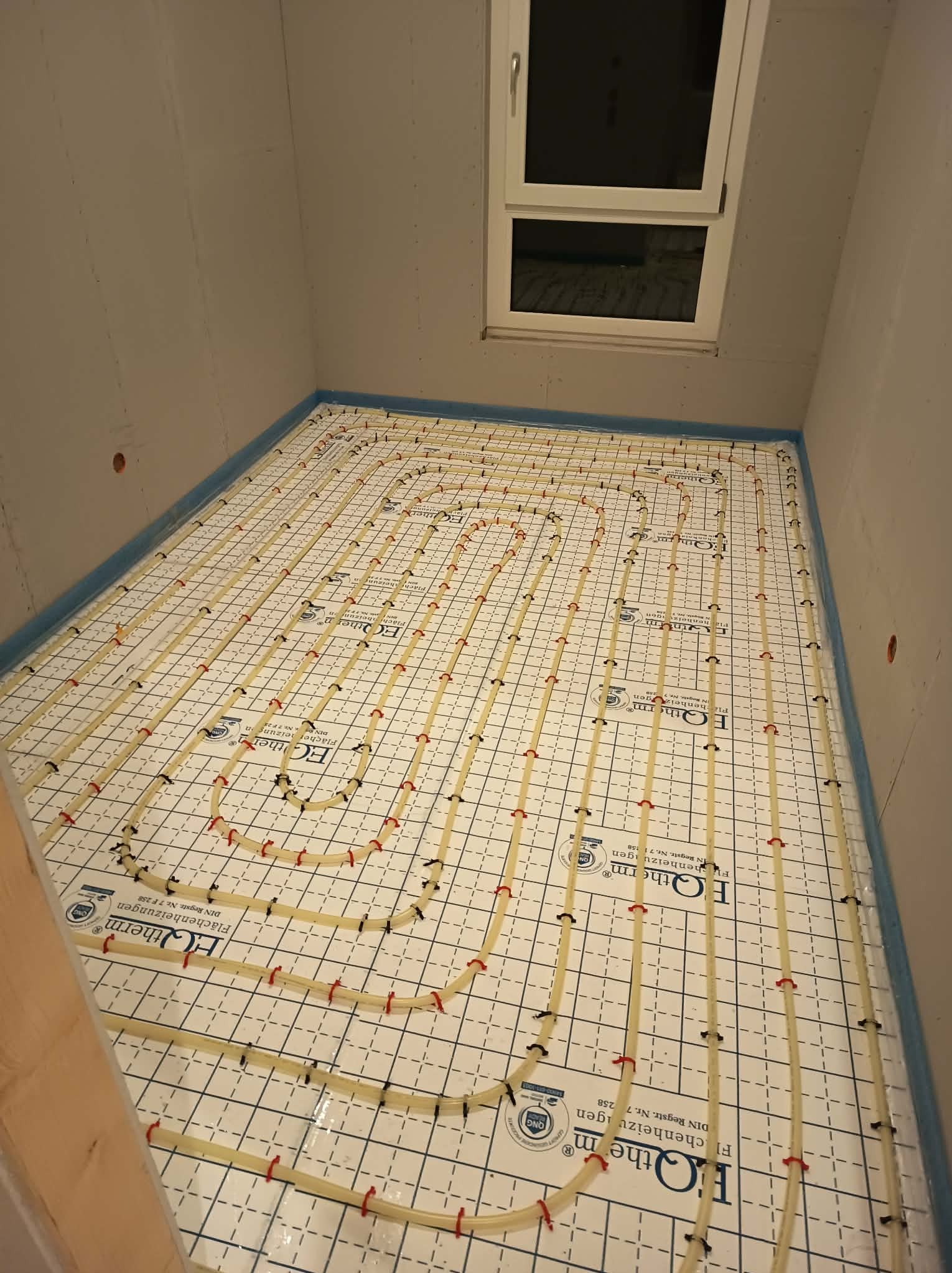 Biała mata grzewcza z rurkami w kształcie pętli, zamocowanymi czerwonymi i czarnymi uchwytami, ułożona na podłodze w pomieszczeniu z oknem dachowym, przygotowana do wylewki.