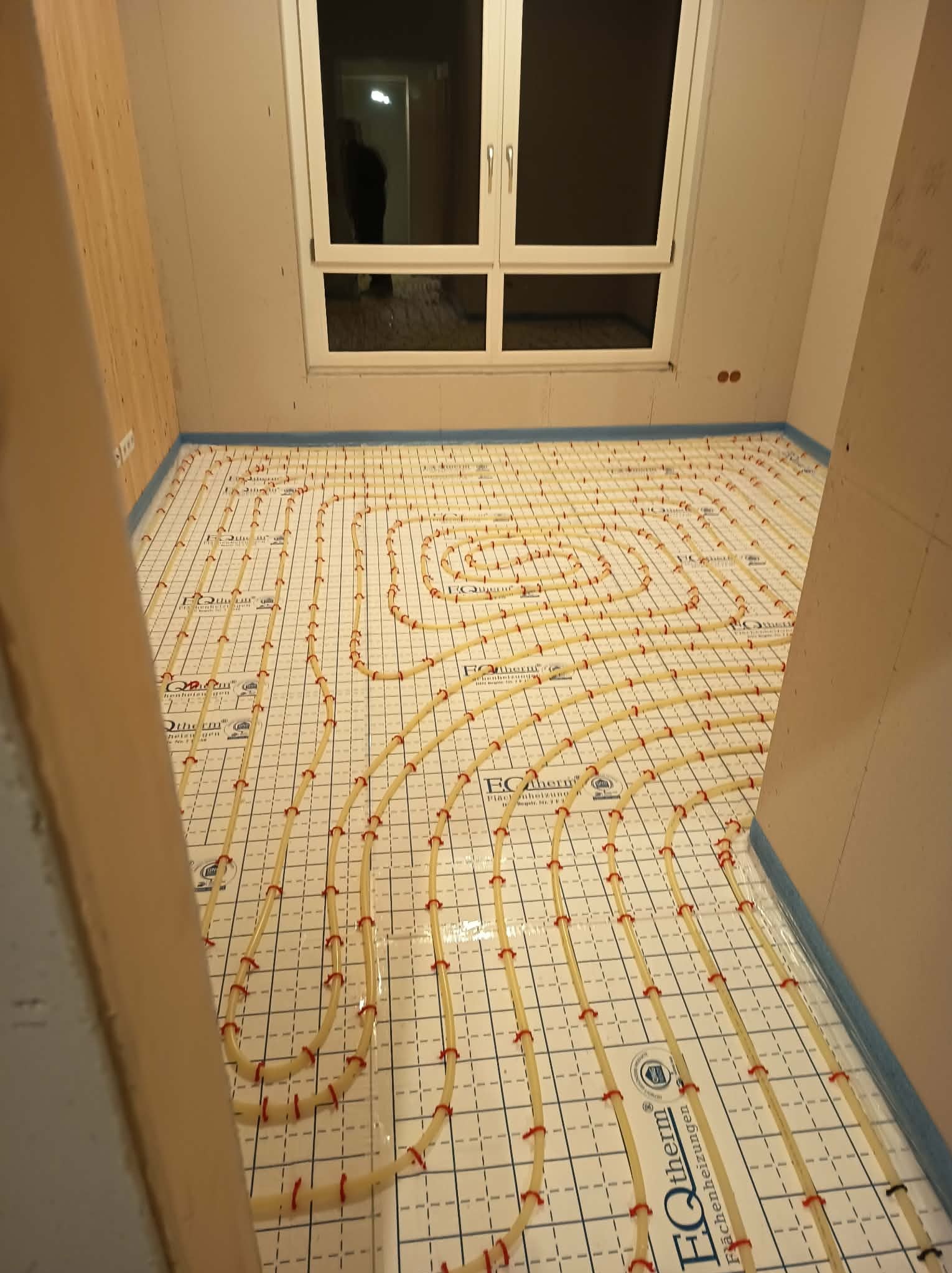 Biała mata grzewcza podłogowa z żółtymi rurkami i czerwonymi uchwytami, ułożona w spiralny wzór w pomieszczeniu z oknem. Widoczny fragment ściany z boazerią.