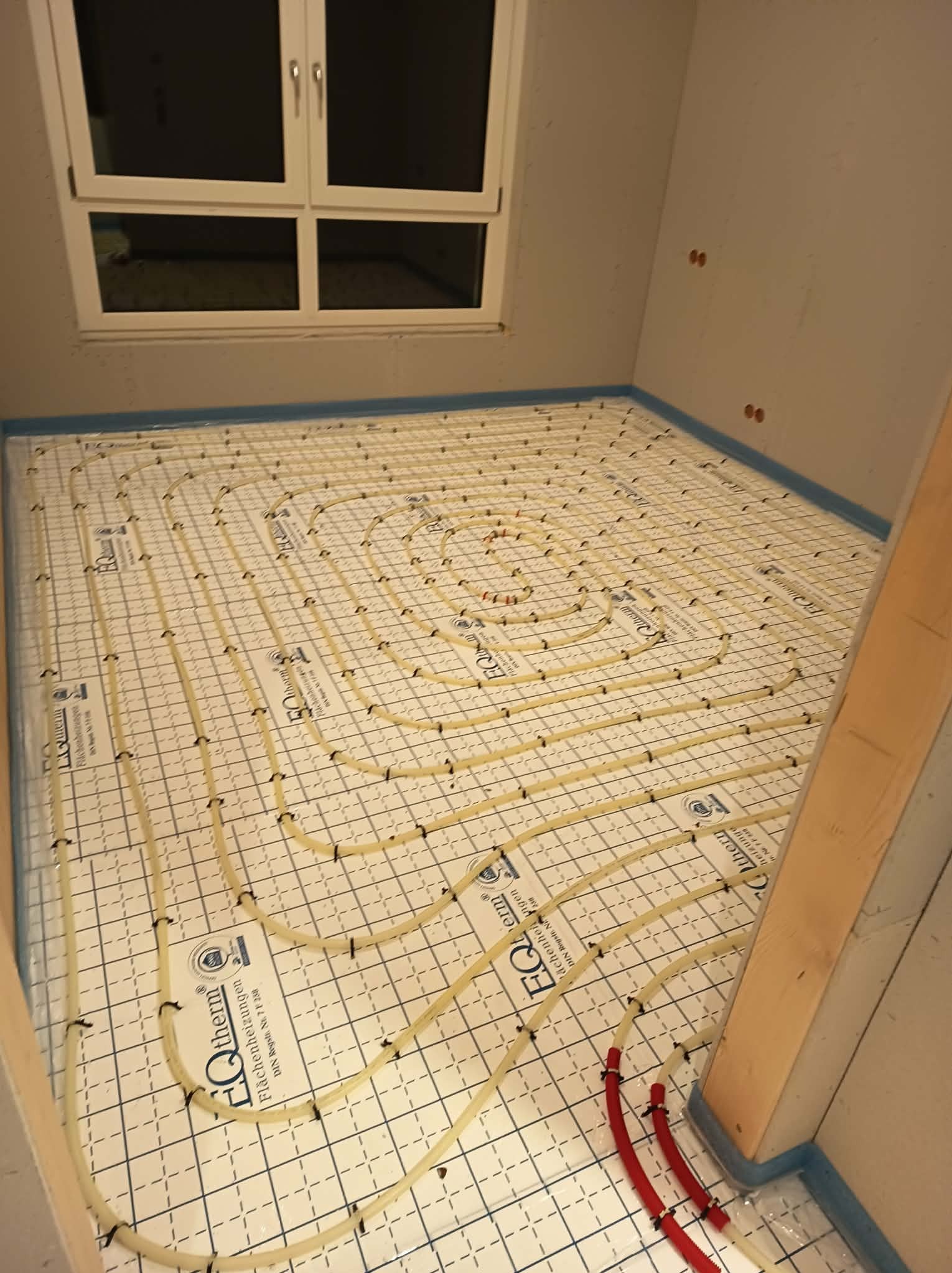 Instalacja ogrzewania podłogowego w łazience, rury ułożone spiralnie na macie z siatką, widoczne przyłącza i okno w tle. Etap prac przed wylaniem posadzki.