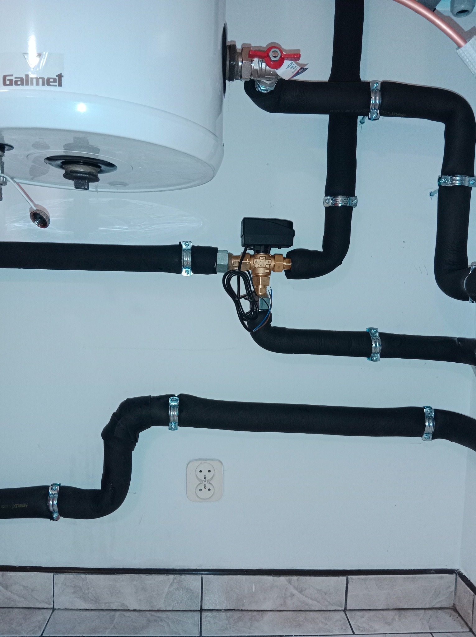 Zamontowany bojler Galmet z izolowanymi rurami i zaworem trójdrożnym na białej ścianie z gniazdkiem elektrycznym i szarą glazurą podłogową. Instalacja wodna w łazience.