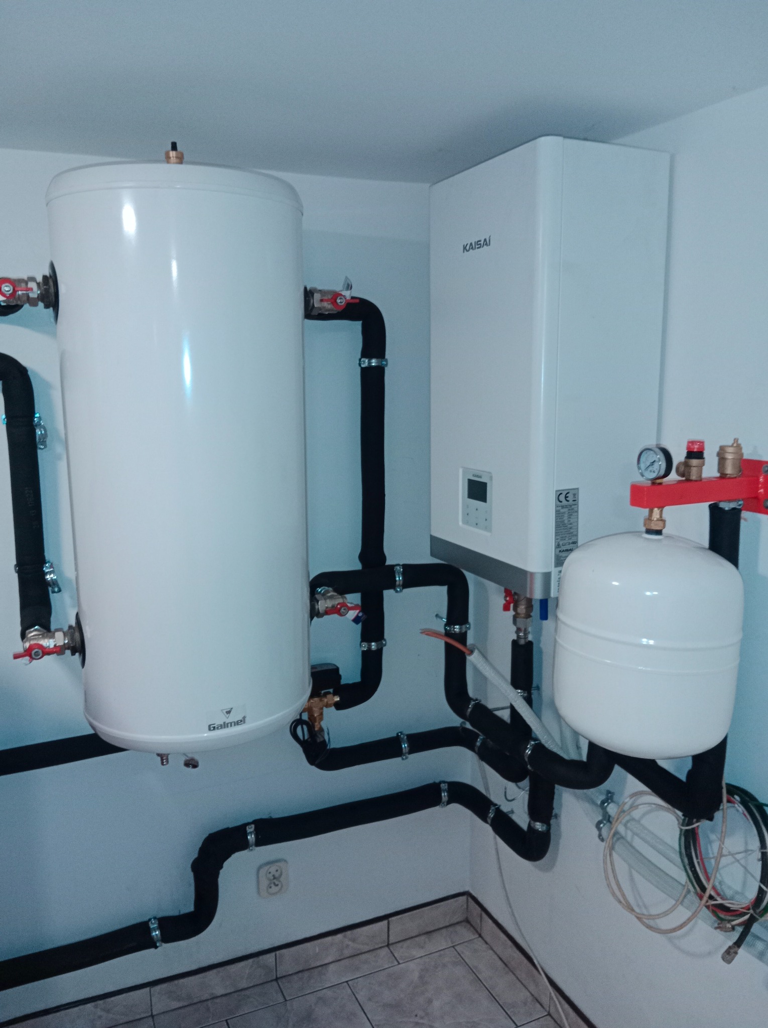 Biały bojler Galmet i piec Kaisai z czarnymi rurami w izolacji, zamontowane na ścianie w pomieszczeniu z szarą podłogą z płytek. Widoczne zawory i manometr.