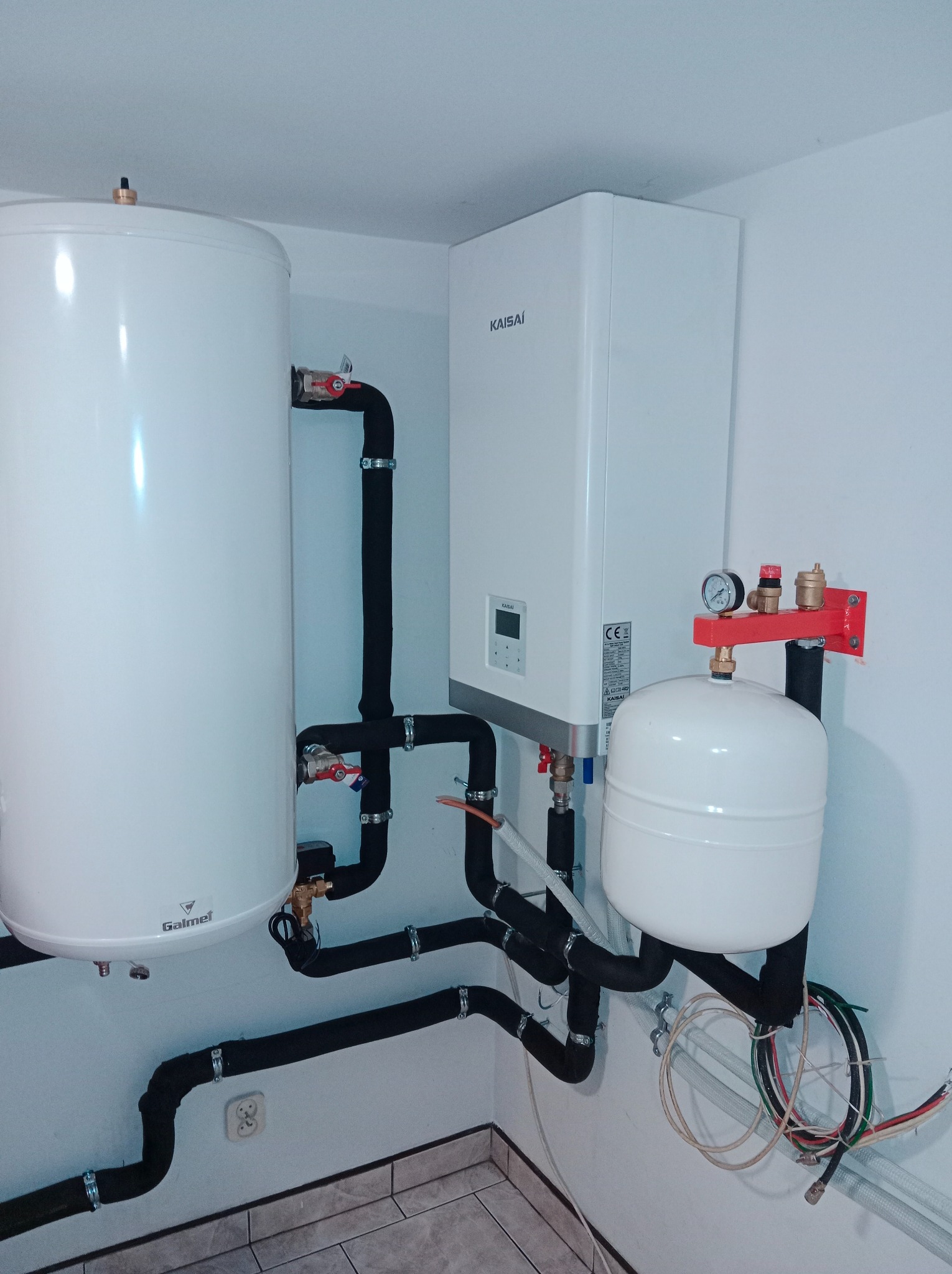 Biały bojler Galmet i piec Kaisai z czarnymi rurami w izolacji, zamontowane na ścianie w pomieszczeniu z szarą podłogą. Widoczne zawory i manometr.