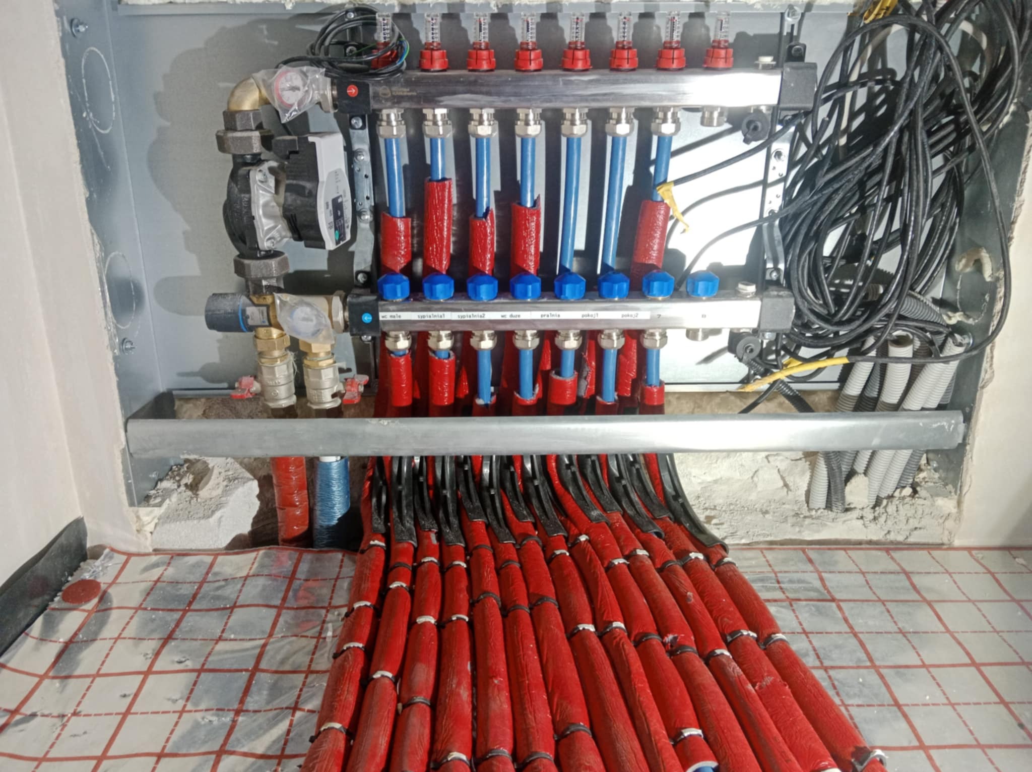 Rozdzielacz ogrzewania podłogowego z czerwonymi rurami na folii grzewczej, widoczne elementy instalacji i okablowanie. Etap prac instalacyjnych w pomieszczeniu.