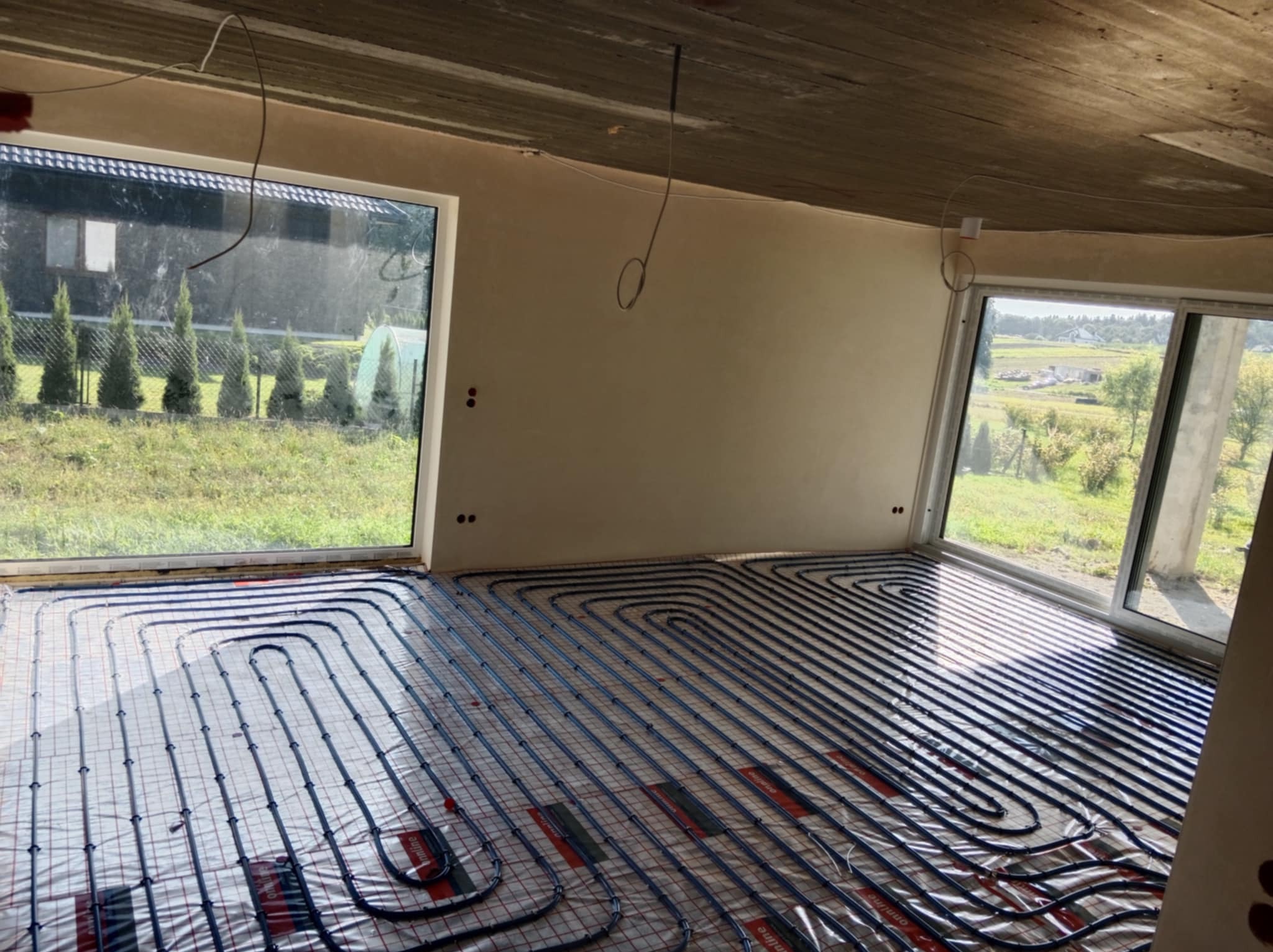 Instalacja ogrzewania podłogowego w surowym wnętrzu z dużymi oknami, widok na zewnątrz. Rury ułożone na folii, ściany przygotowane do dalszych prac wykończeniowych.