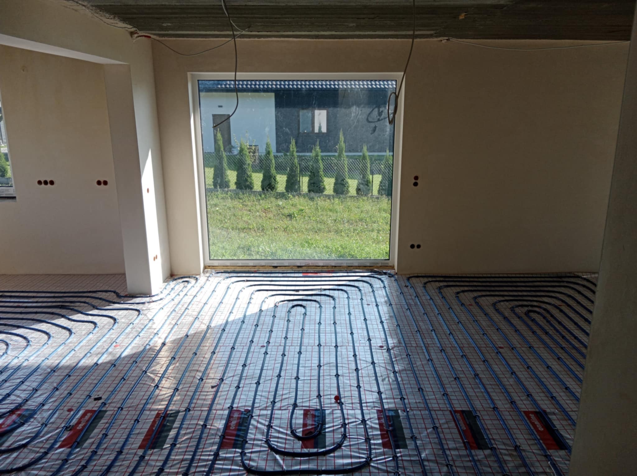 Instalacja ogrzewania podłogowego w surowym wnętrzu z widokiem na zieleń przez okno. Równomiernie rozłożone rury na folii, przygotowane do zalania posadzką.