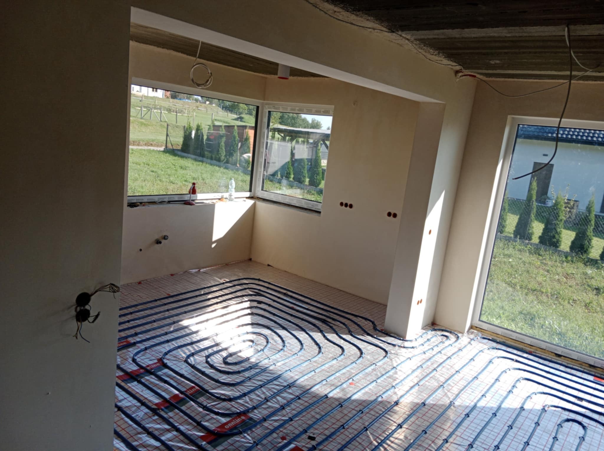 Instalacja ogrzewania podłogowego w jasnym pomieszczeniu z dużymi oknami, widok na zewnątrz. Rury ułożone na folii izolacyjnej, ściany przygotowane do dalszych prac wykończeniowych.