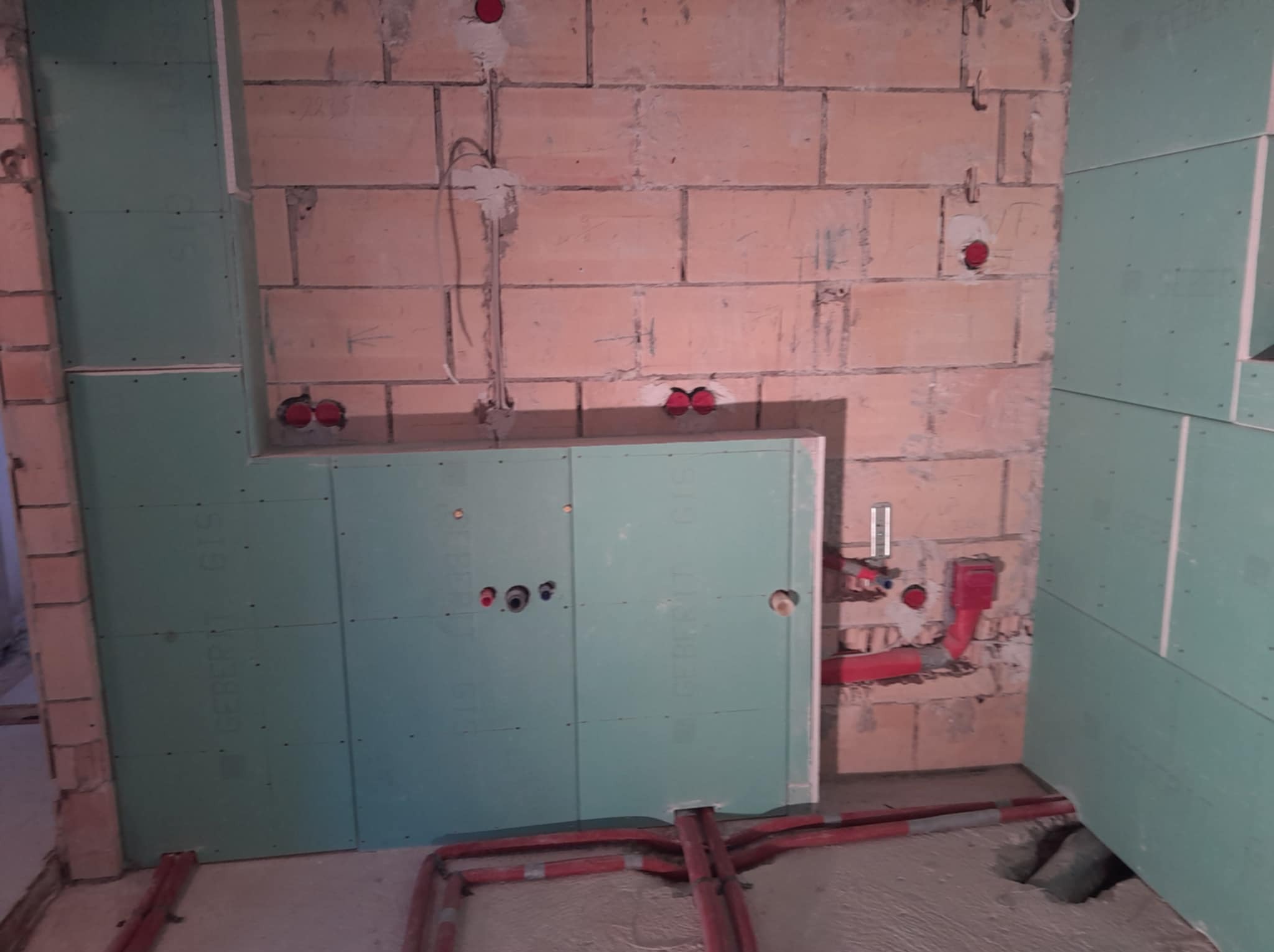 Łazienka w trakcie remontu: ściany z cegły i płyty gipsowej, widoczne instalacje wodne i elektryczne. Etap prac przed wykończeniem.