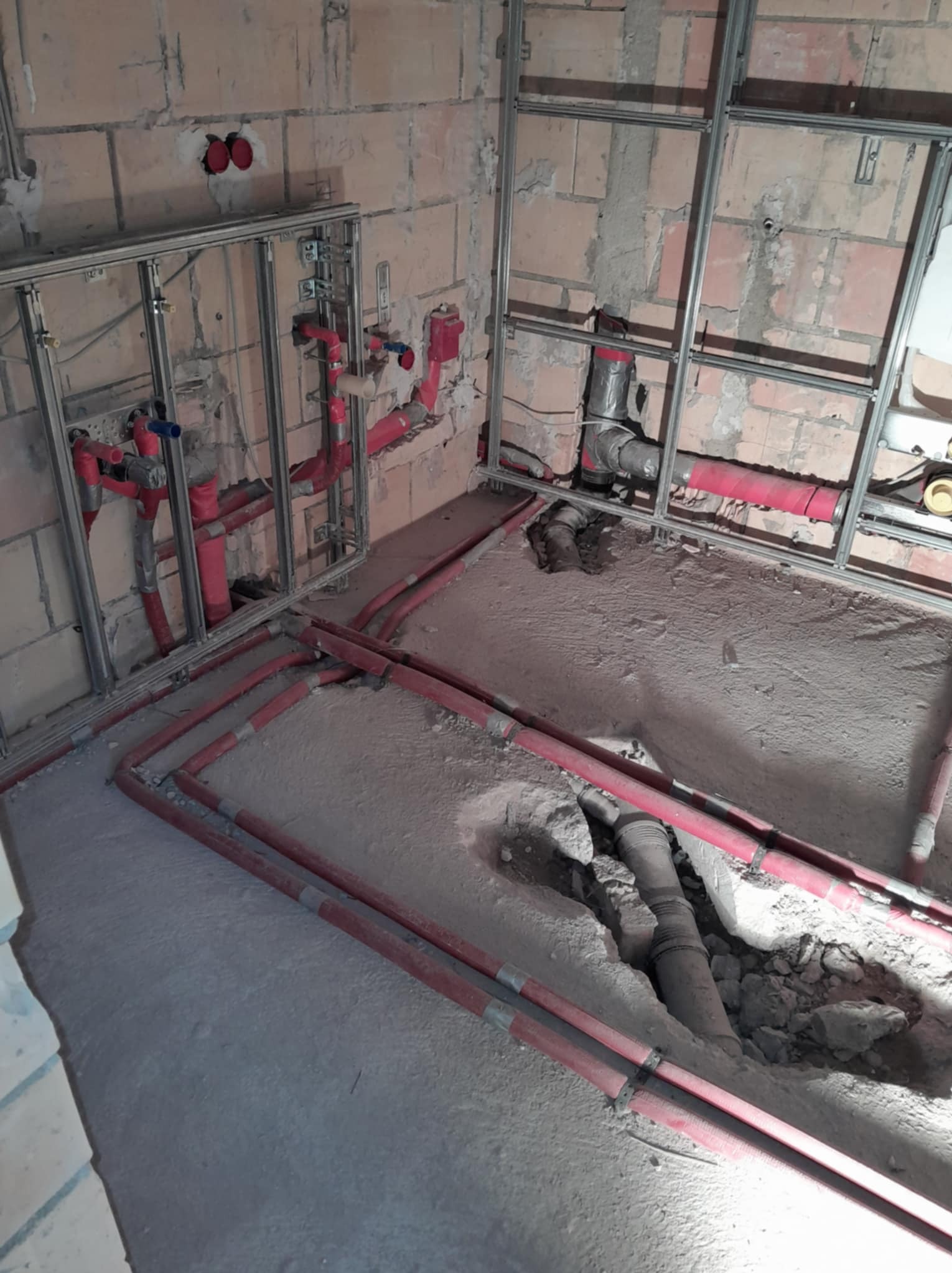 Instalacja wodno-kanalizacyjna w trakcie montażu: czerwone rury prowadzone wzdłuż ścian i podłogi, widoczne metalowe profile konstrukcyjne na tle surowej cegły.