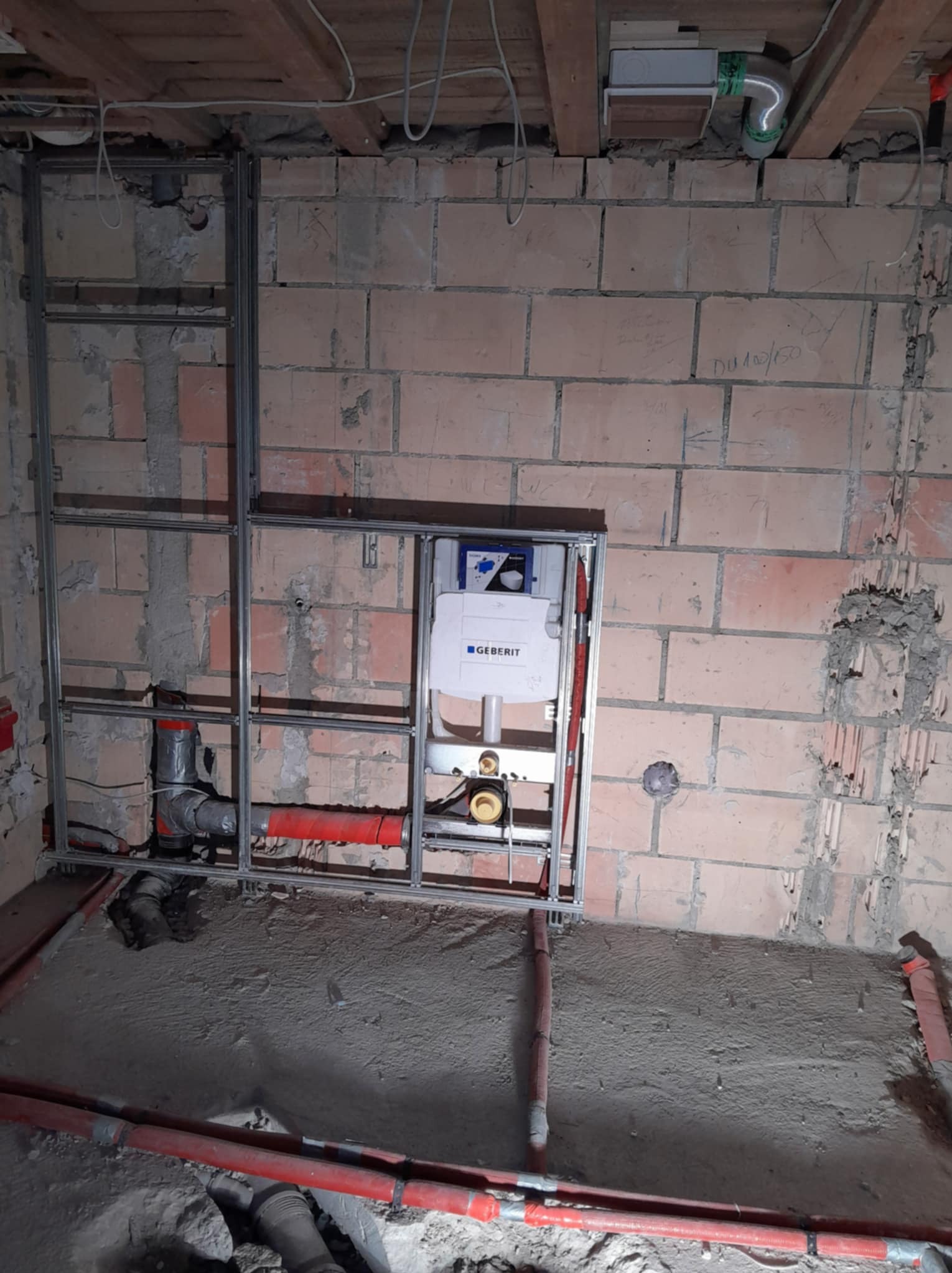 Montaż stelaża podtynkowego Geberit w surowej łazience z widocznymi instalacjami wodno-kanalizacyjnymi i metalową konstrukcją. Ściany z cegły, podłoga wylewka.