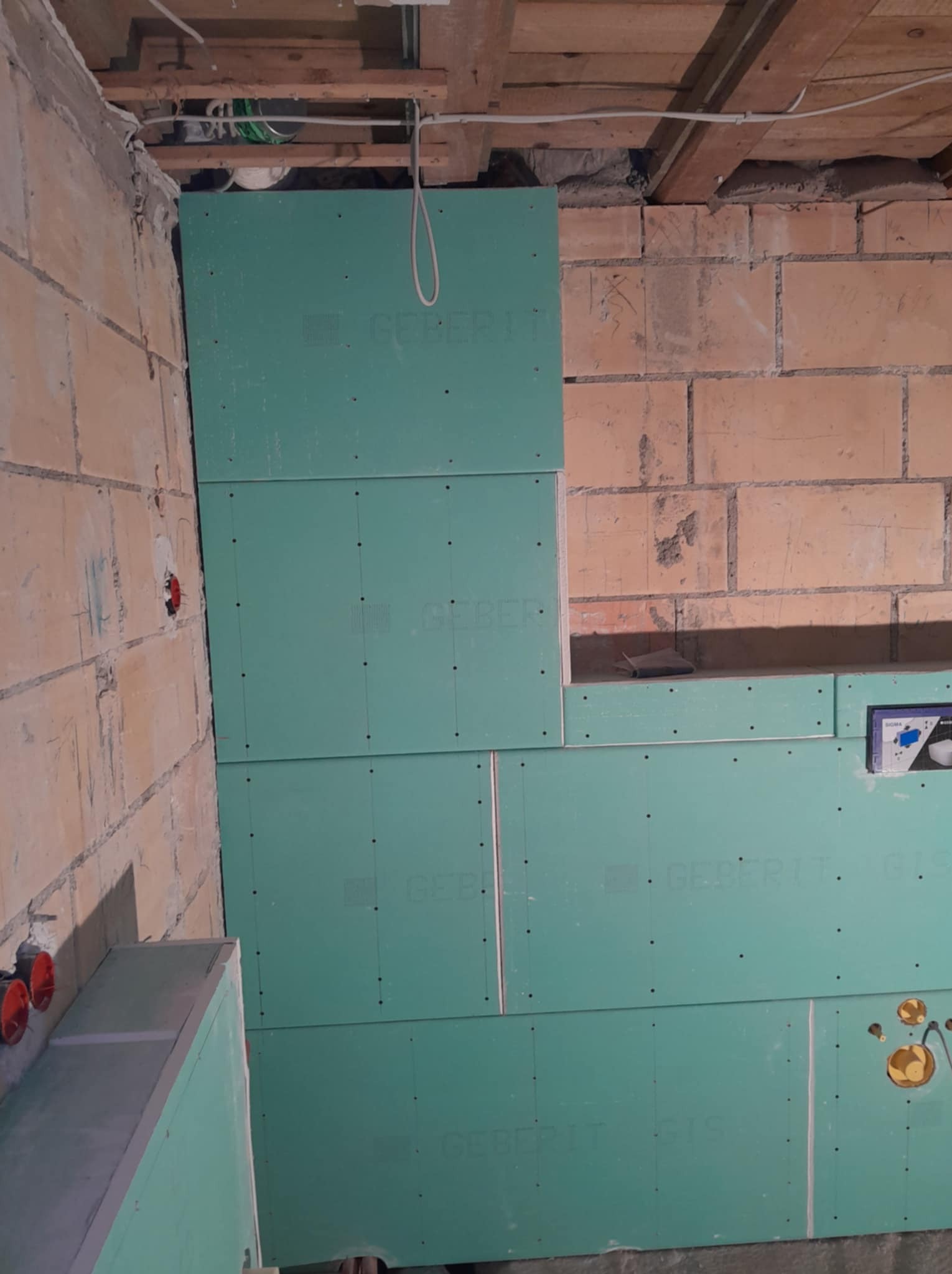 Montaż płyt gipsowo-kartonowych Geberit na ścianie z cegły w łazience. Widoczne otwory na instalacje i fragment sufitu z drewnianymi belkami. Etap prac wykończeniowych.