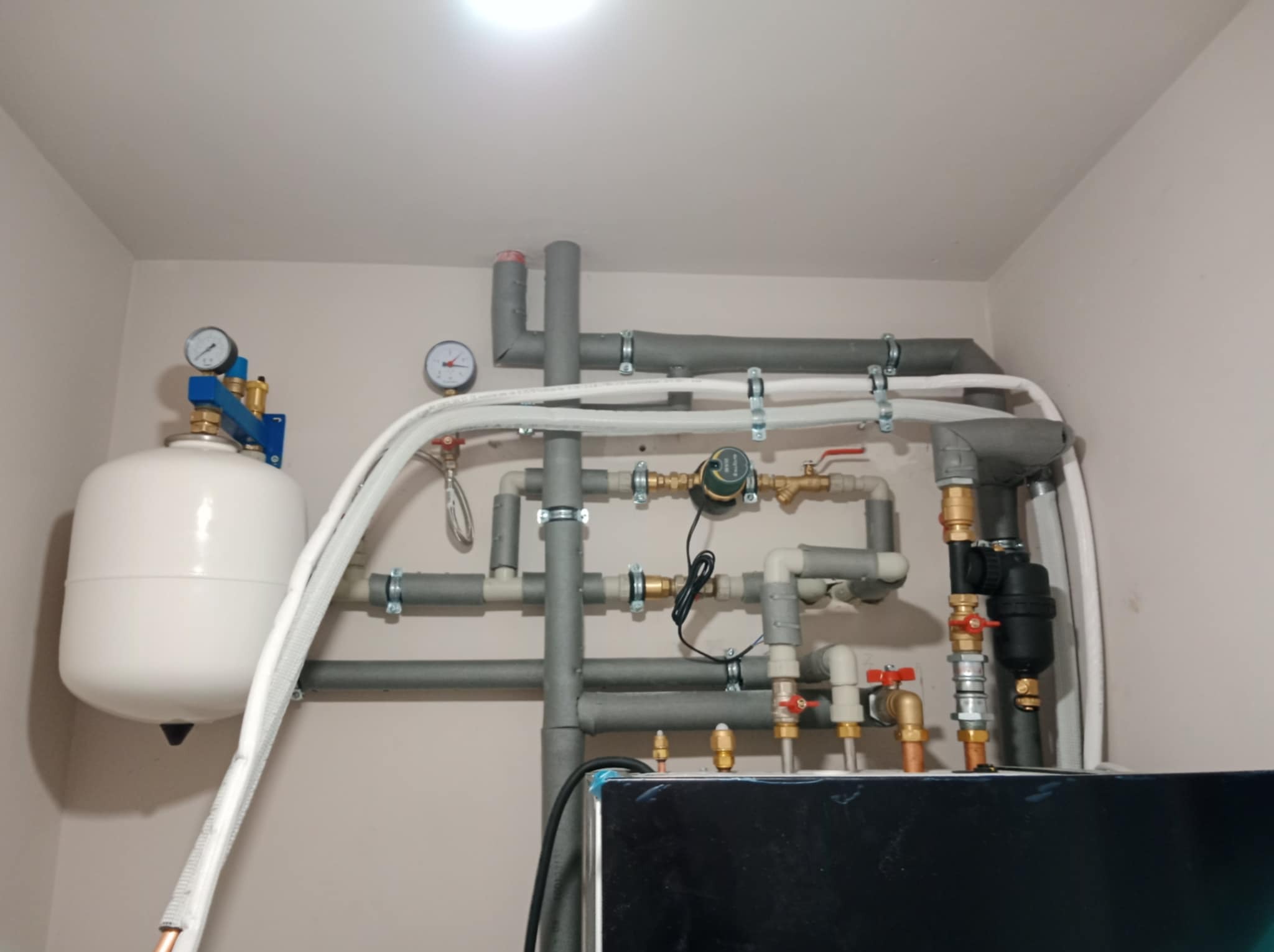 Instalacja grzewcza z szarymi rurami, manometrami i białym zbiornikiem wyrównawczym zamontowana na ścianie w pomieszczeniu. Widoczne zawory i izolacja termiczna.