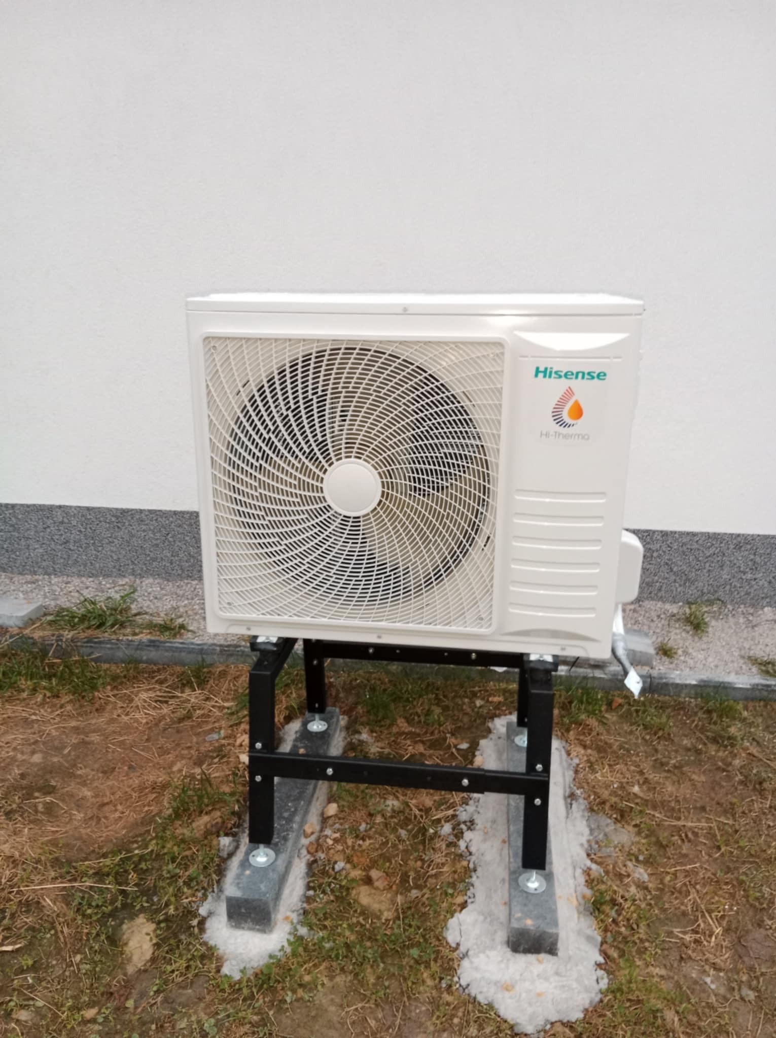 Biała jednostka zewnętrzna klimatyzacji Hisense na czarnym stelażu, umieszczona na betonowych fundamentach przy białej ścianie budynku. Widoczna trawa i kostka brukowa.