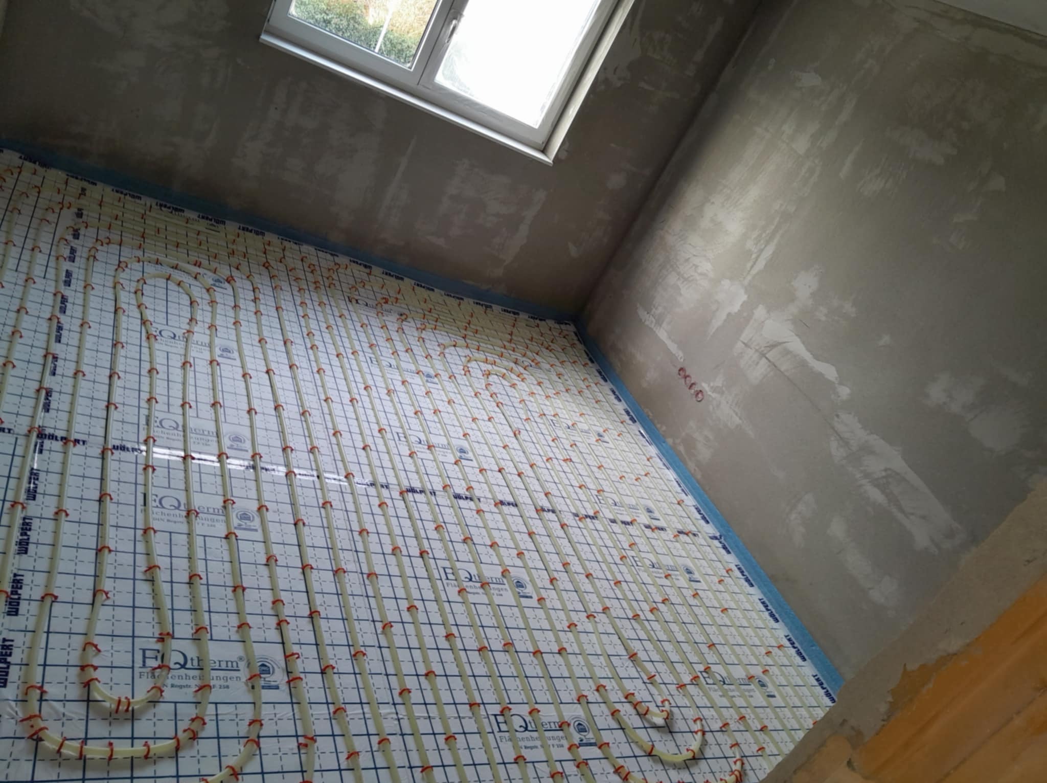 Instalacja ogrzewania podłogowego na folii z siatką w surowym wnętrzu łazienki, widok z góry, okno dachowe w tle. Etap prac wykończeniowych.