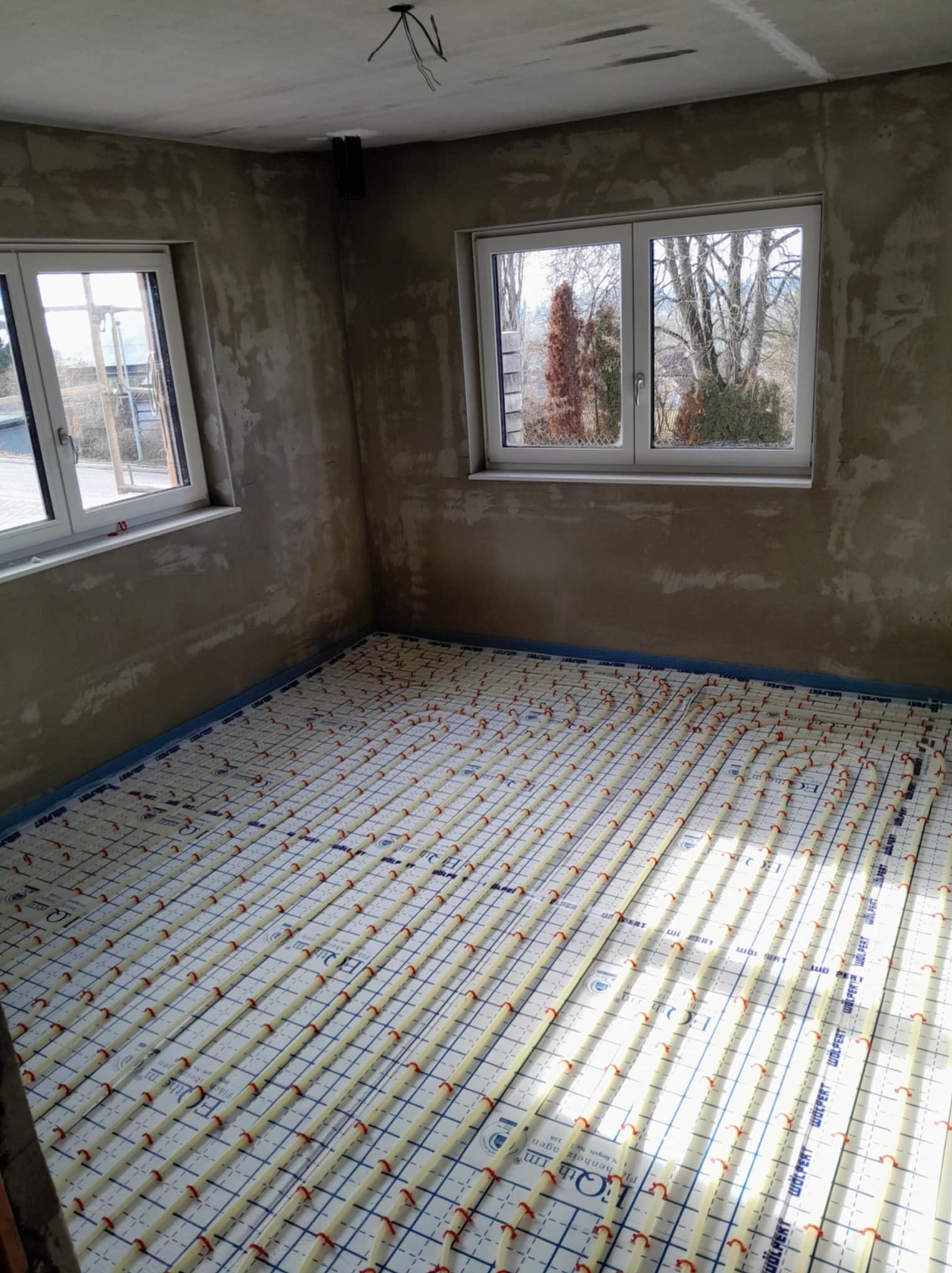 Instalacja ogrzewania podłogowego w surowym wnętrzu. Widoczne rury grzewcze na izolacji, ściany w stanie deweloperskim, dwa okna z widokiem na zewnątrz.