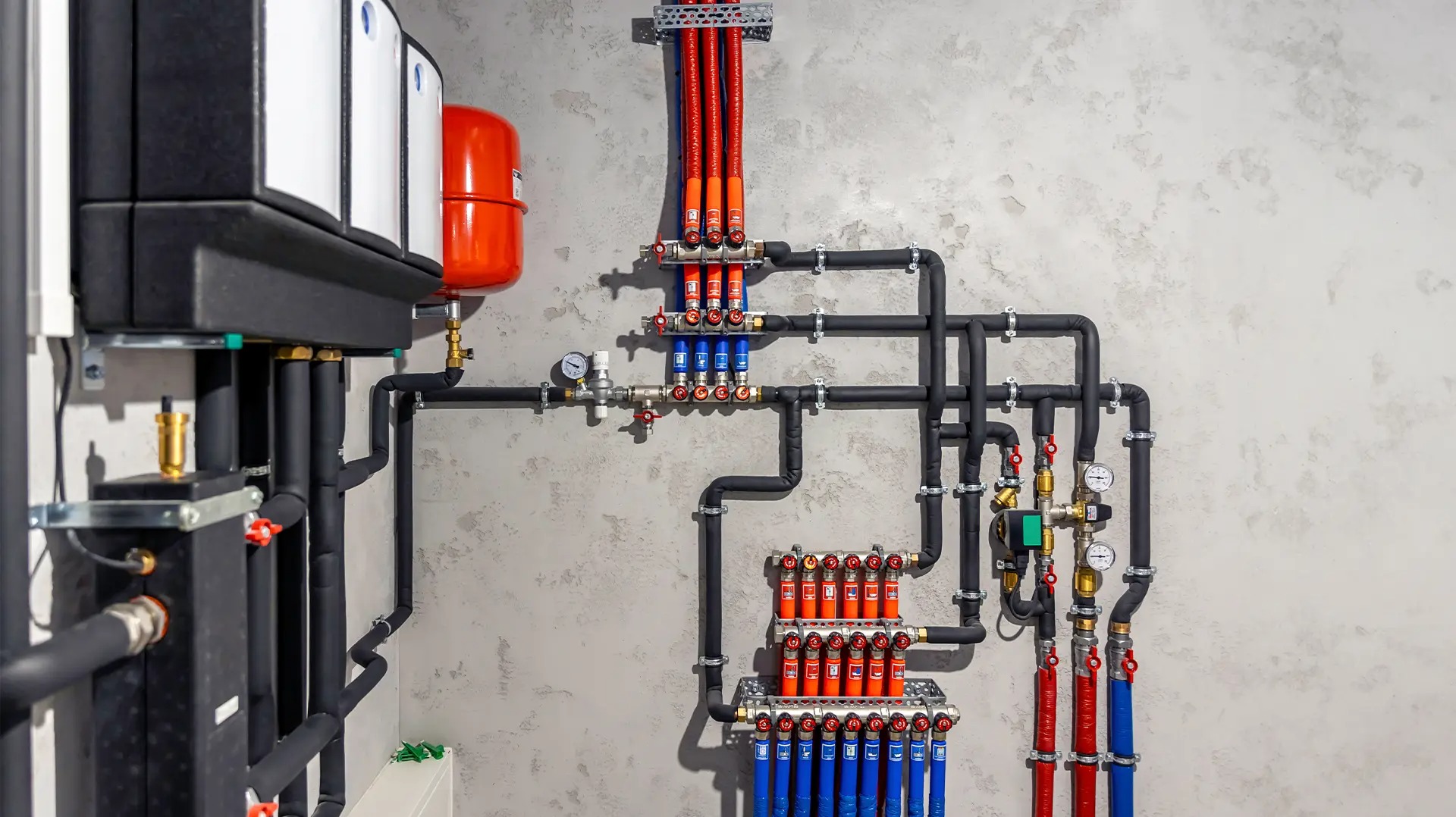 Złożona instalacja grzewcza z czarnymi rurami, czerwonymi i niebieskimi przewodami, manometrami i zaworami na tle szarej ściany o nieregularnej fakturze.