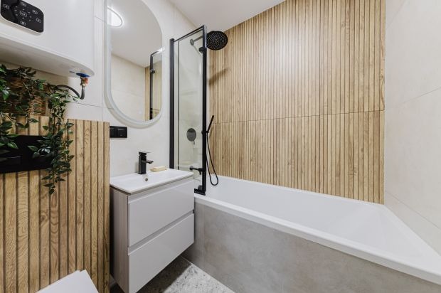 Nowoczesna łazienka z wanną, kabiną prysznicową z czarnymi elementami, umywalką z szafką i drewnianą okładziną ścienną. Minimalistyczny design, jasne kolory.