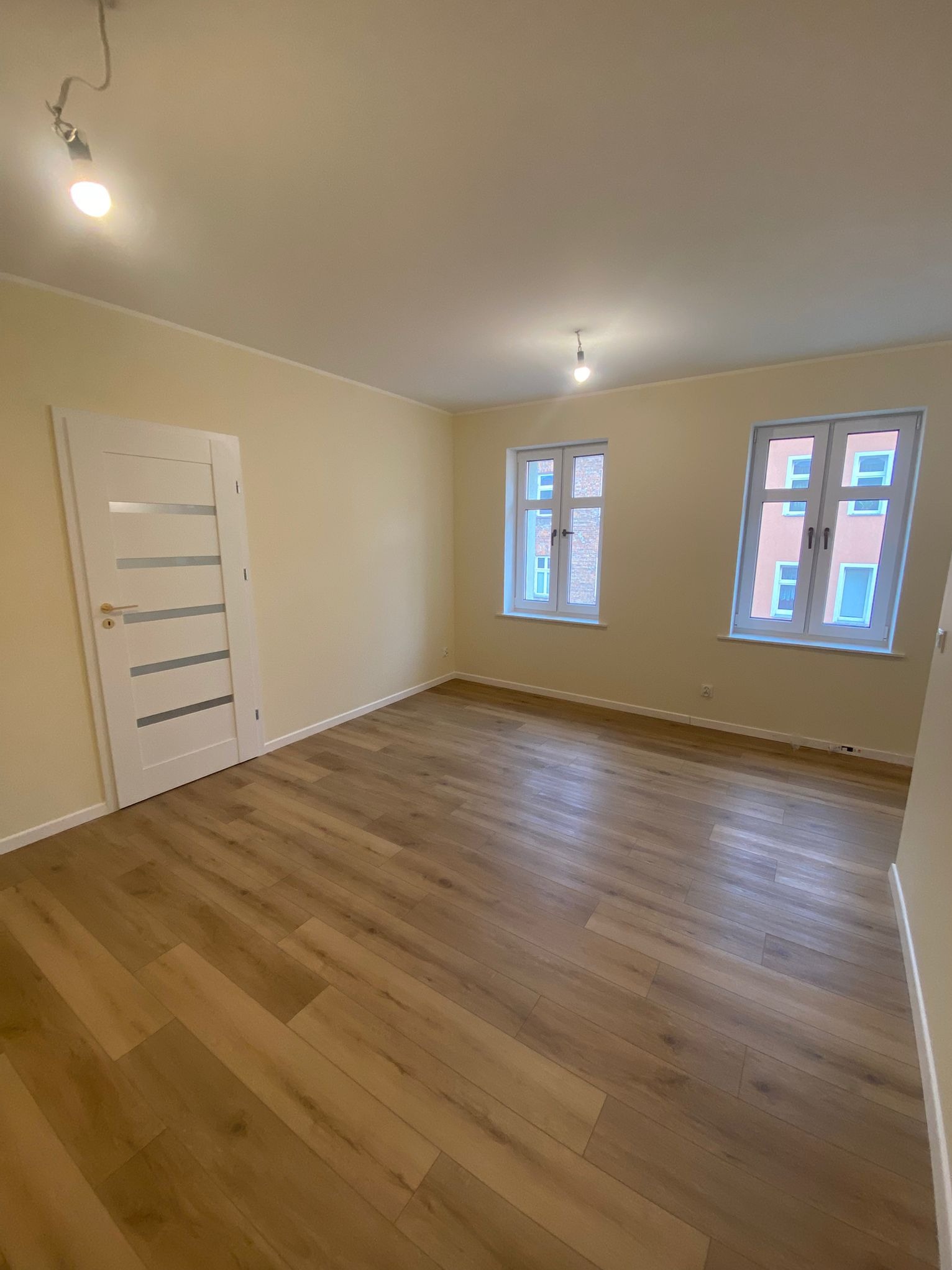 Jasne wnętrze po remoncie: parkiet, pomalowane ściany na kremowo, białe okna i drzwi z przeszkleniami. Widok na dwa okna i drzwi z boku.