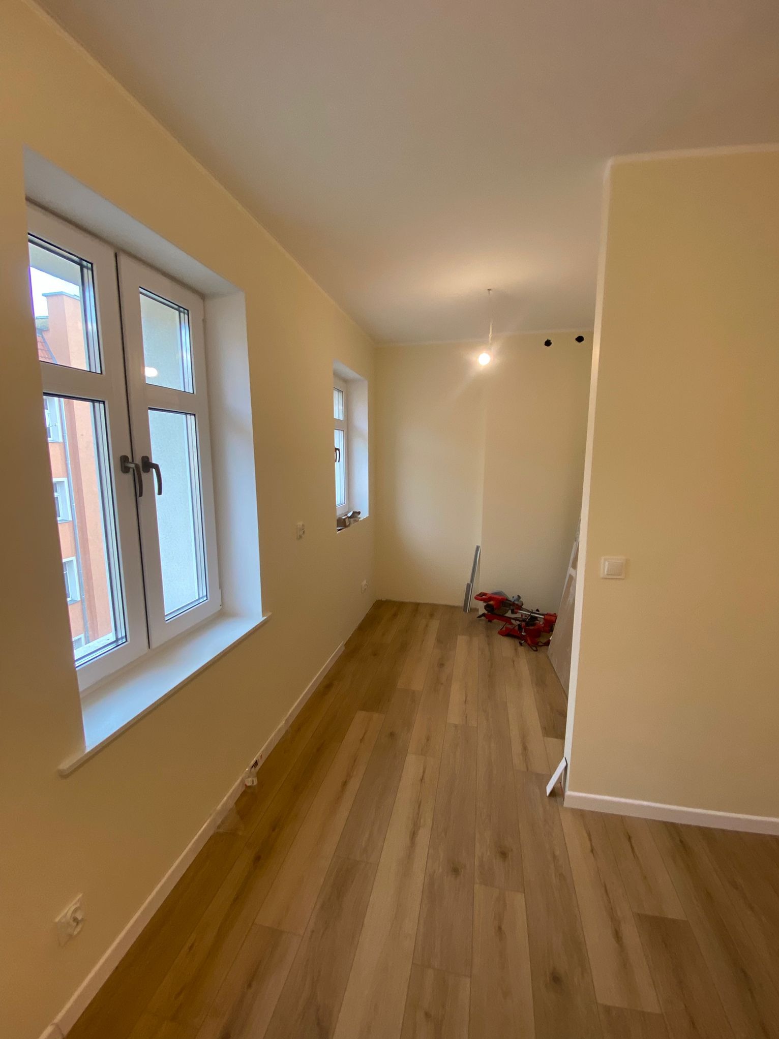 Jasne wnętrze po remoncie: drewniana podłoga, świeżo pomalowane ściany w odcieniach beżu, białe okna. W głębi widoczne narzędzia i drabina.