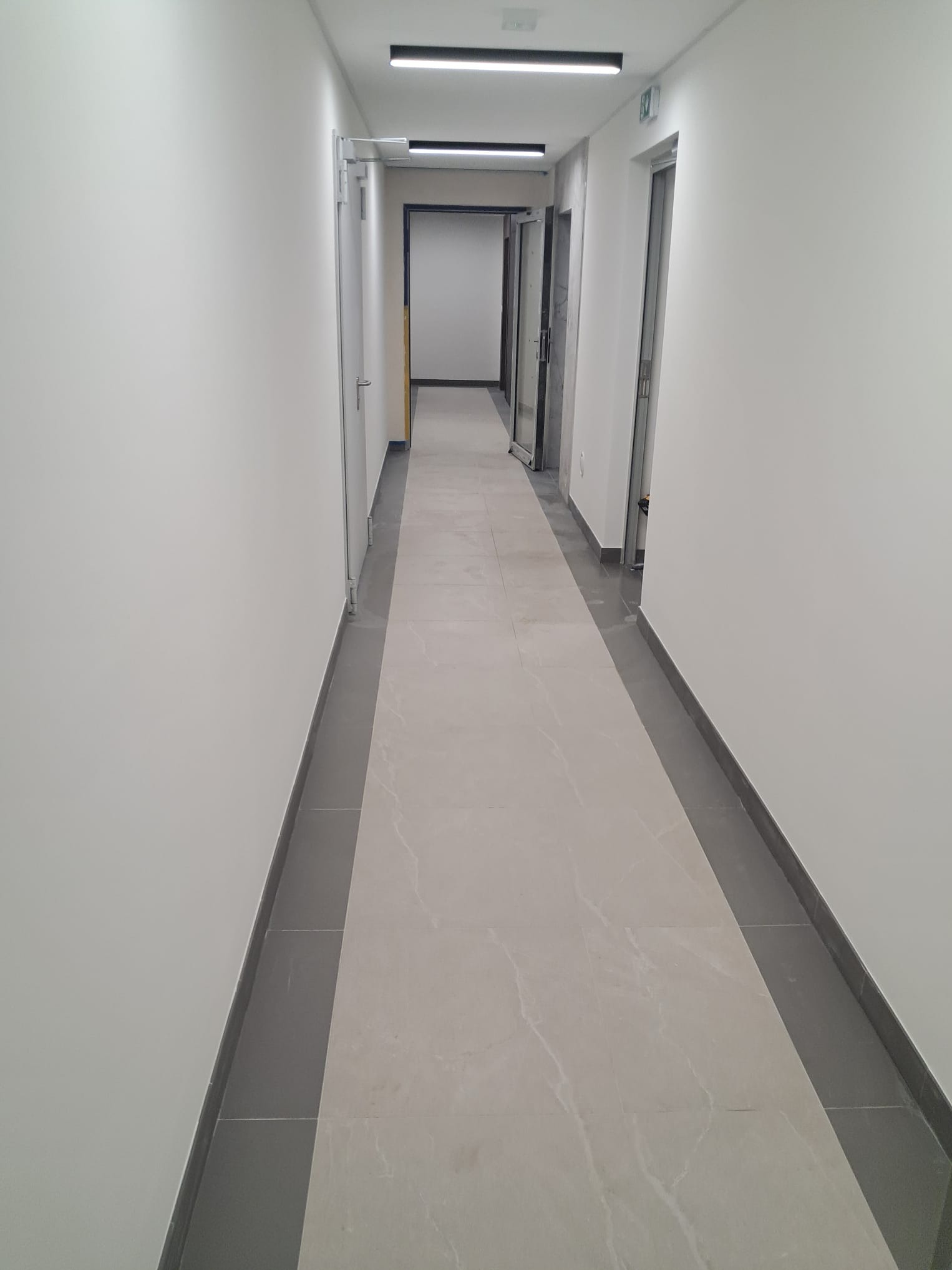 Wykończony korytarz biurowy z jasnymi ścianami, szarą podłogą z płytek i nowoczesnym oświetleniem LED. Widok w perspektywie, ukazujący czystość i minimalizm wnętrza.