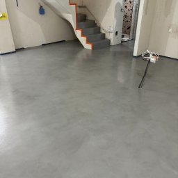 Technodecor Yevhen Syvodiedov - Szara, gładka posadzka w jasnym wnętrzu z białymi schodami o pomarańczowych krawędziach. Widoczne niedokończone prace wykończeniowe i wystające kable.